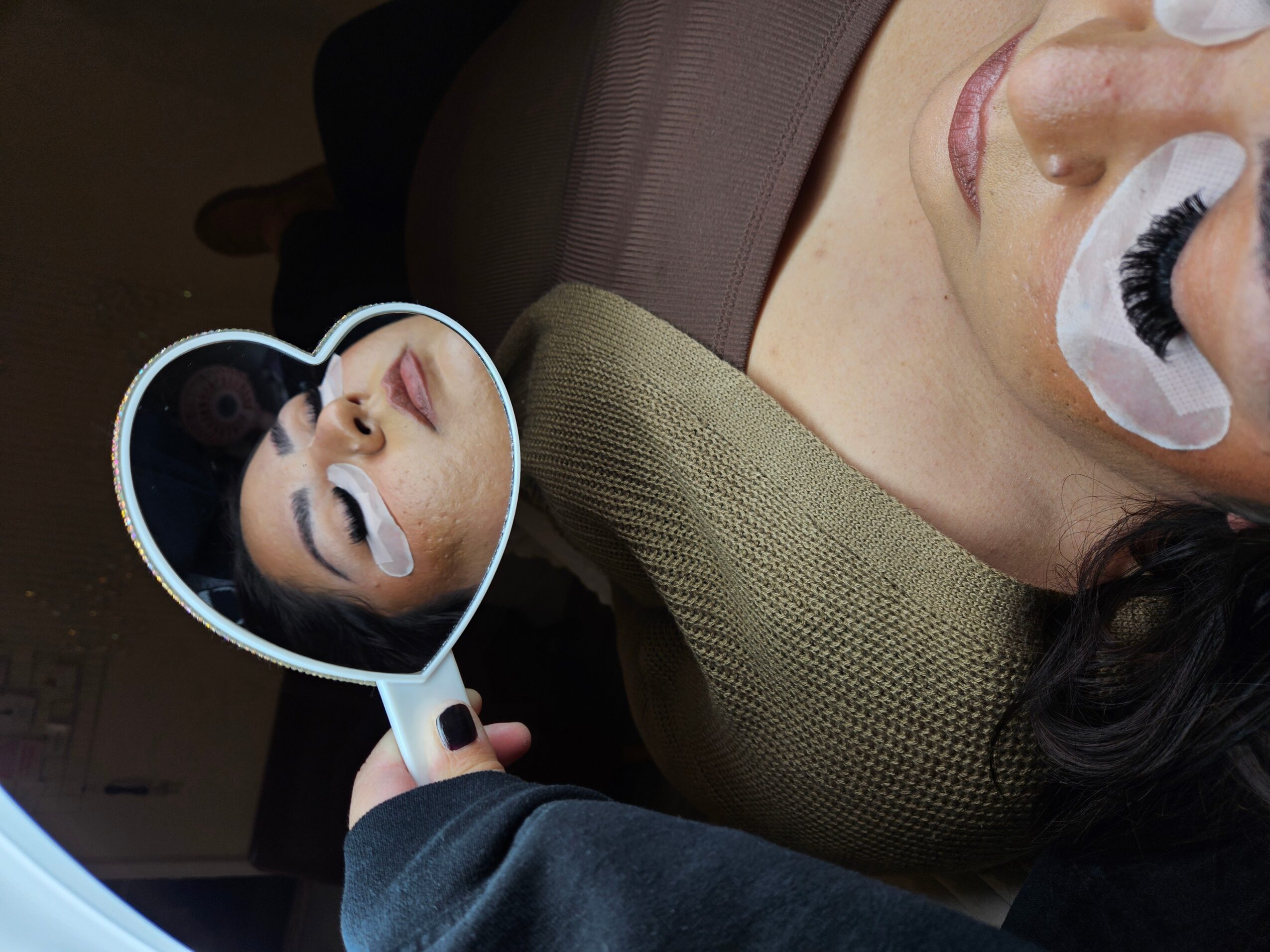 Person lying down with eye patches, looking into a mirror held by another person, showing their face. 20 words.