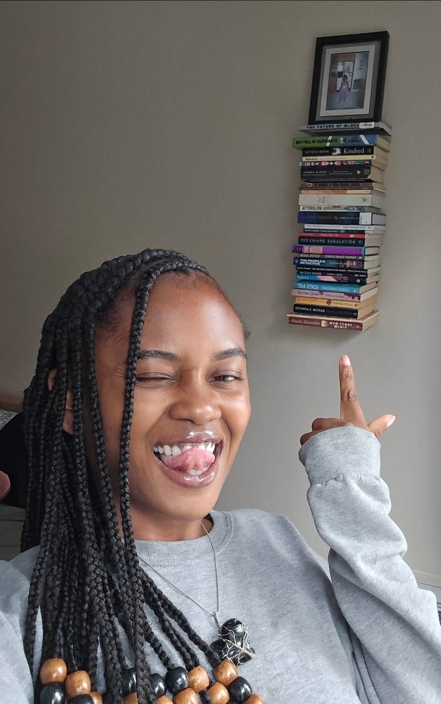 Smiling person with braided hair sticking out tongue, pointing at a tall stack of books on a wall shelf, in a room.
