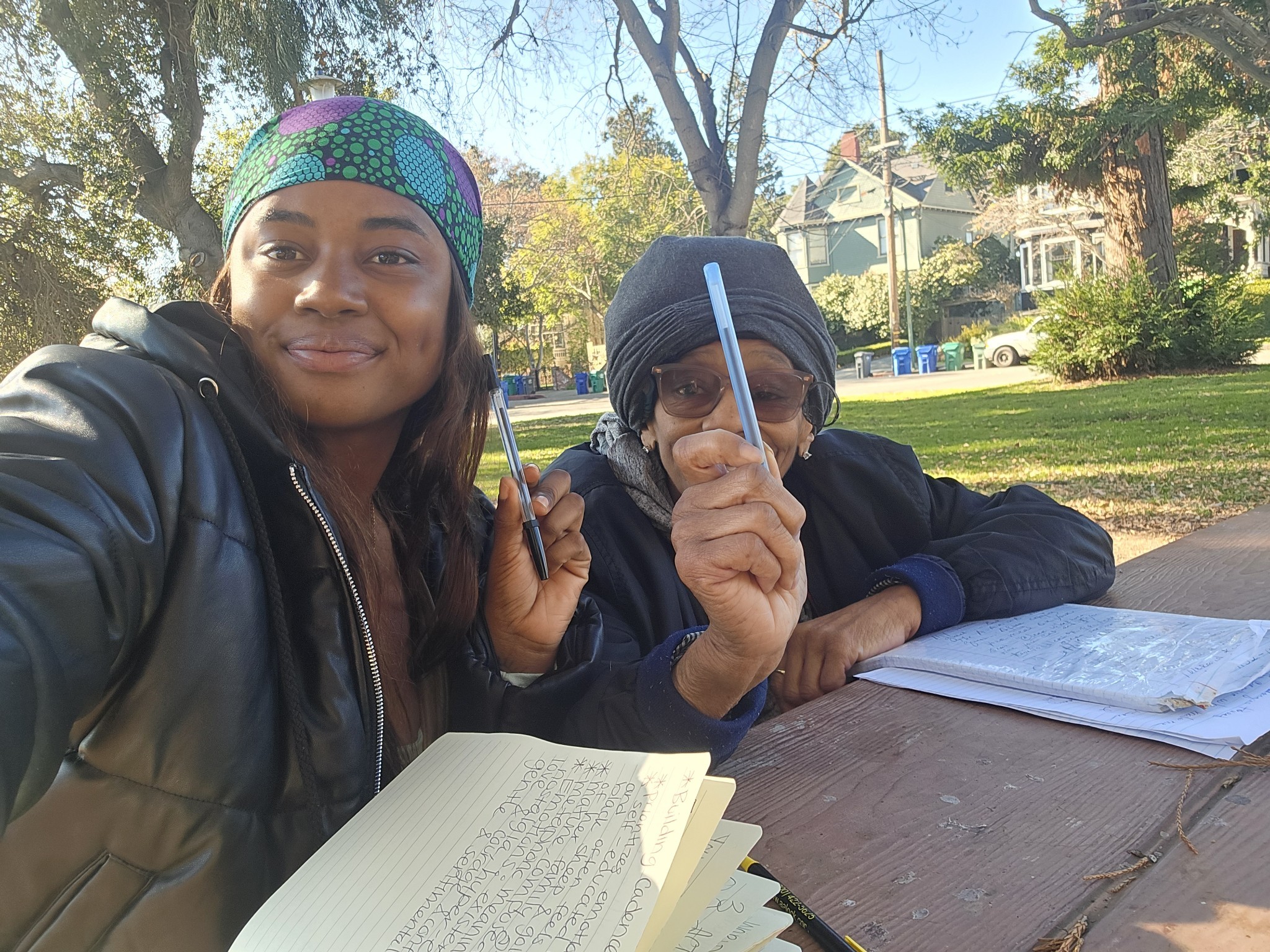 Two people sitting at a table outdoors with notebooks, one smiling and the other holding a pen, trees and houses in background.