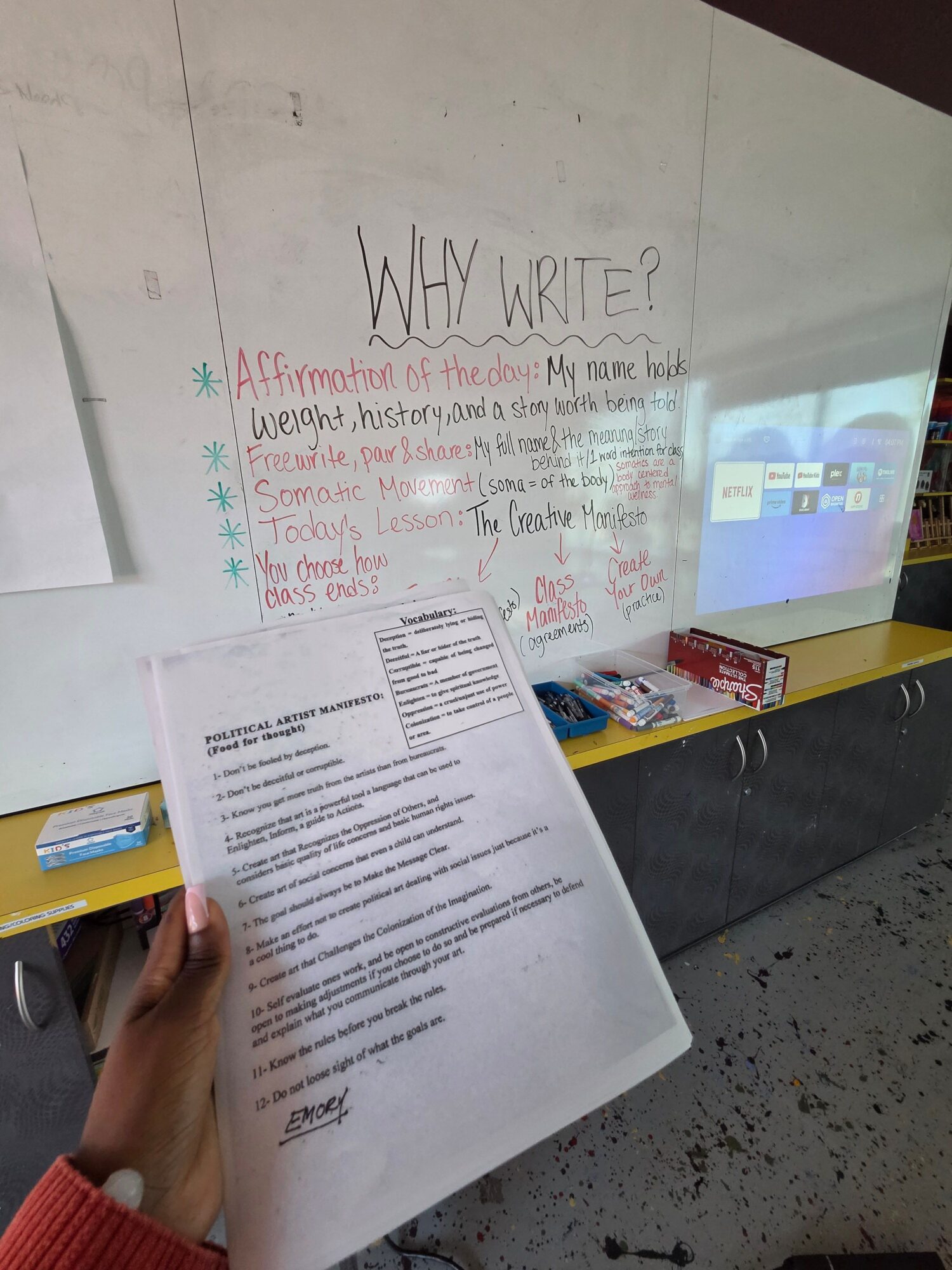 Person holding a paper with printed text, on a table with a whiteboard, markers, and a box of supplies.