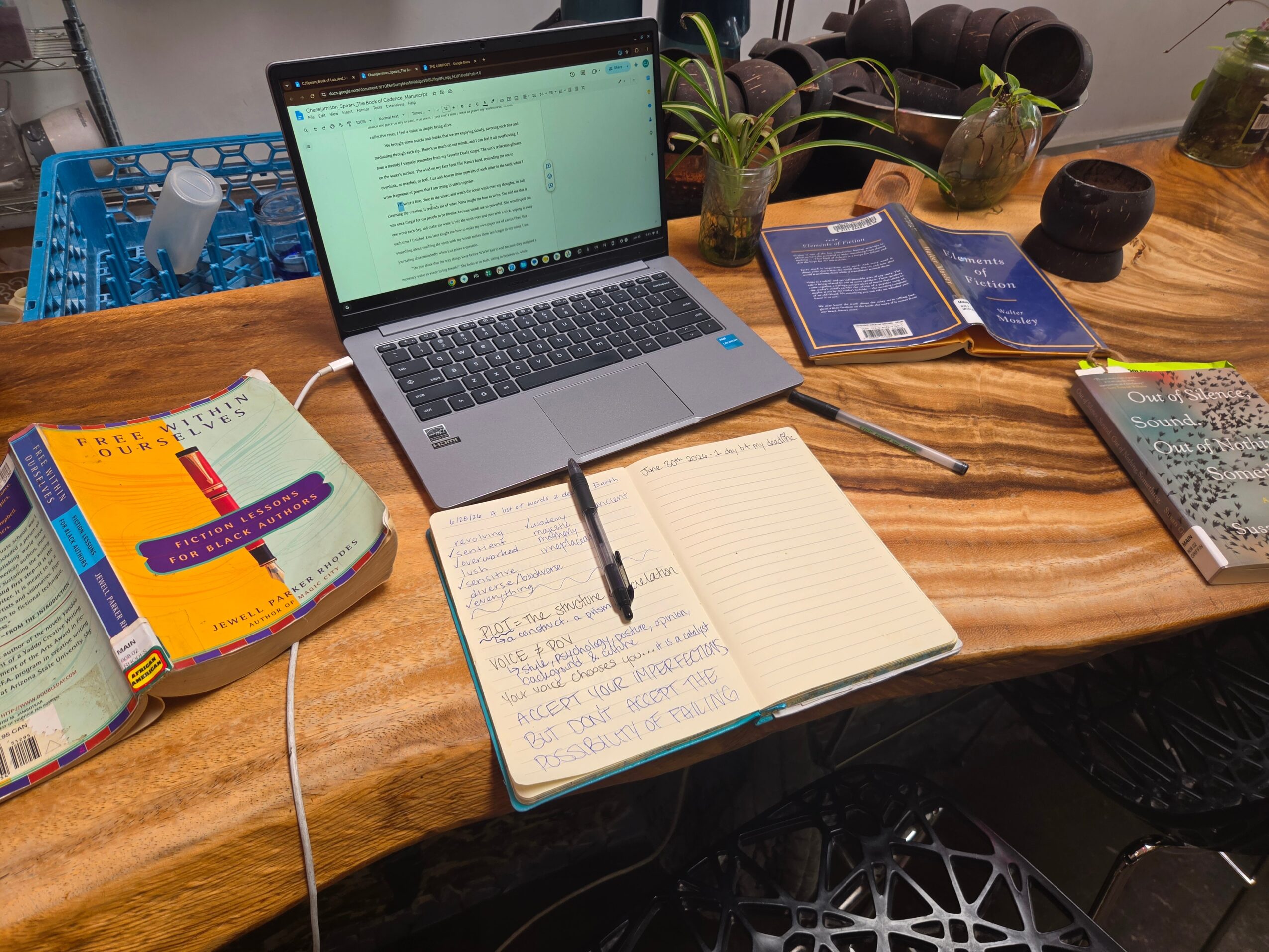 Desk with open laptop, notebooks, pens, books, and plants, arranged on a wooden surface.