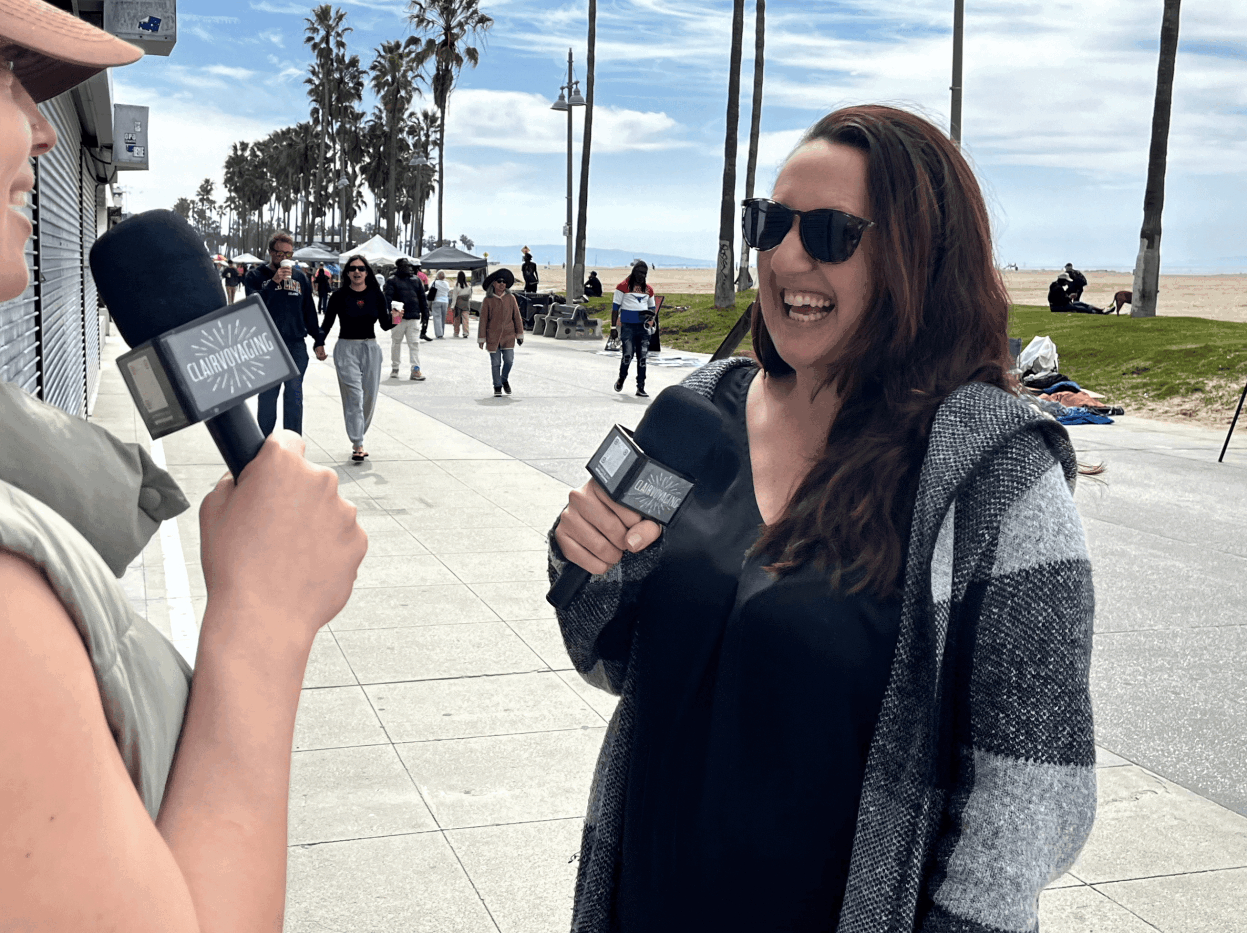 Woman with sunglasses smiling, holding a microphone, outdoors near a beach with palm trees and people in the background.