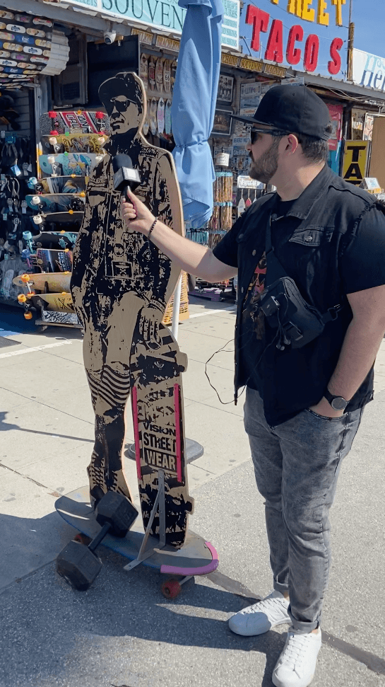 Man in black vest and cap interacts with cardboard cutout of a woman holding a sign, outdoors on a sunny day.