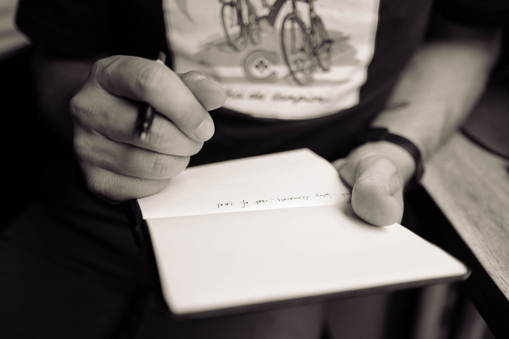 Person writing in a notebook with a pen, wearing a wristwatch and a T-shirt with a bicycle graphic.