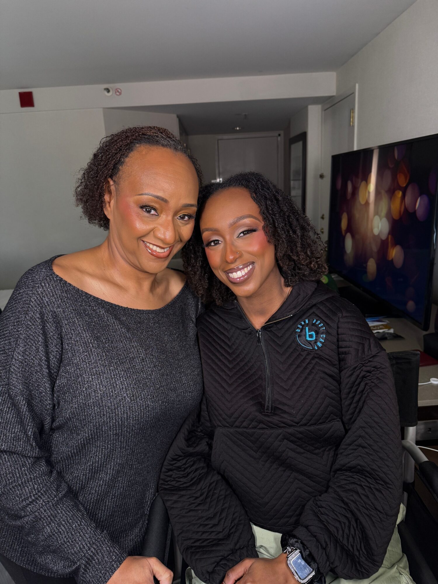 Two women smiling, one with curly hair and the other with straight hair, in an indoor setting.