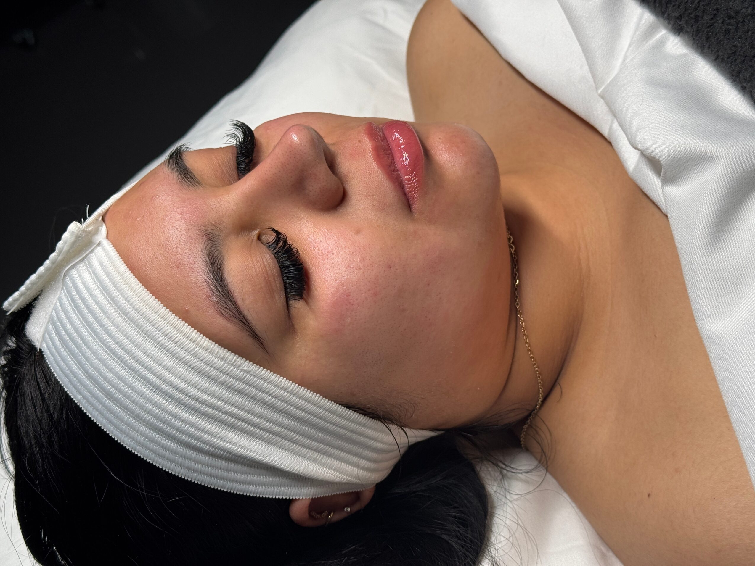 Close-up of a woman with eyes closed, lying down with a white headband, makeup, and a white garment visible.