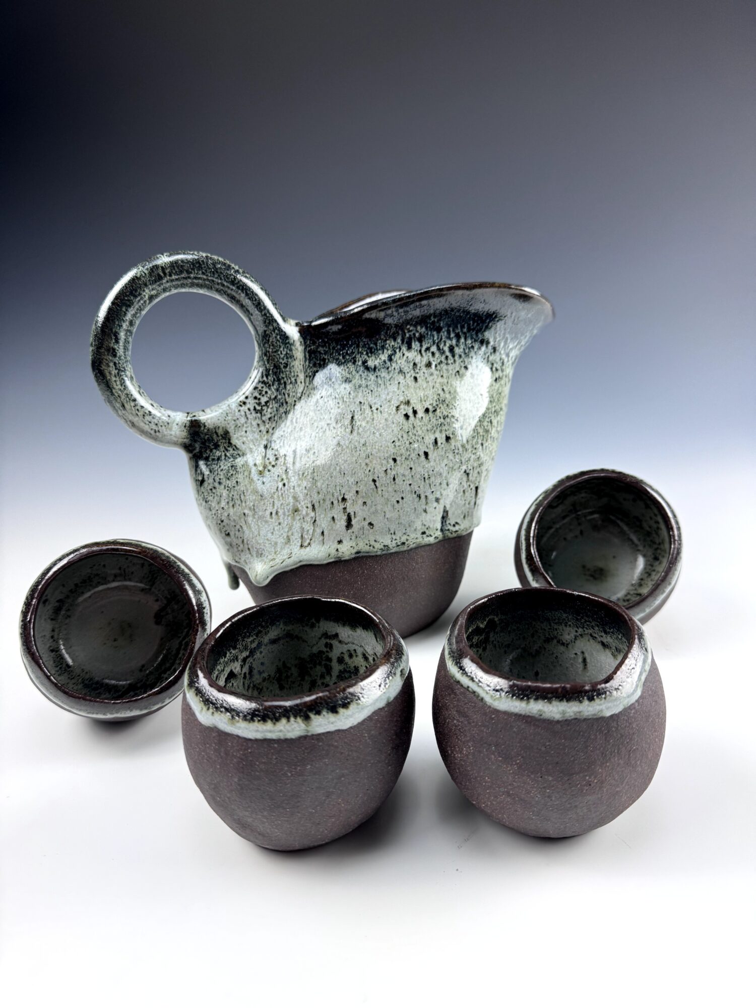 Ceramic pitcher with four matching cups arranged around it, set against a gradient background.