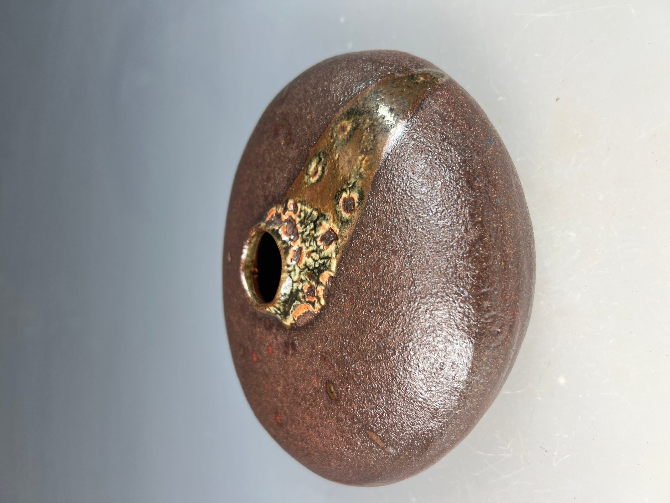 Round object with a metallic patch and a hole, surface appears textured and brownish, against a plain background.