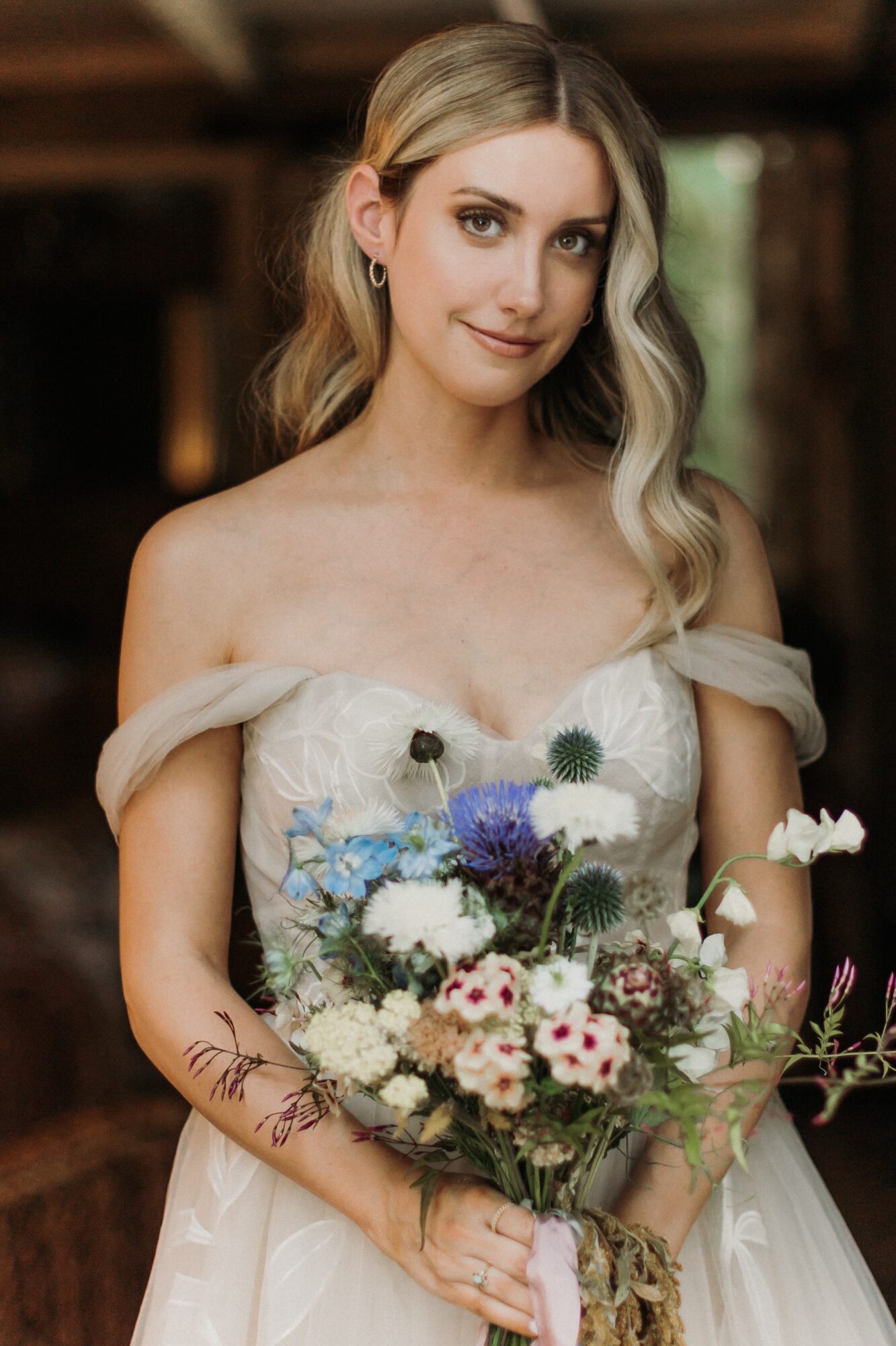 Young woman in wedding dress holding a bouquet of flowers, smiling indoors with blurred background.