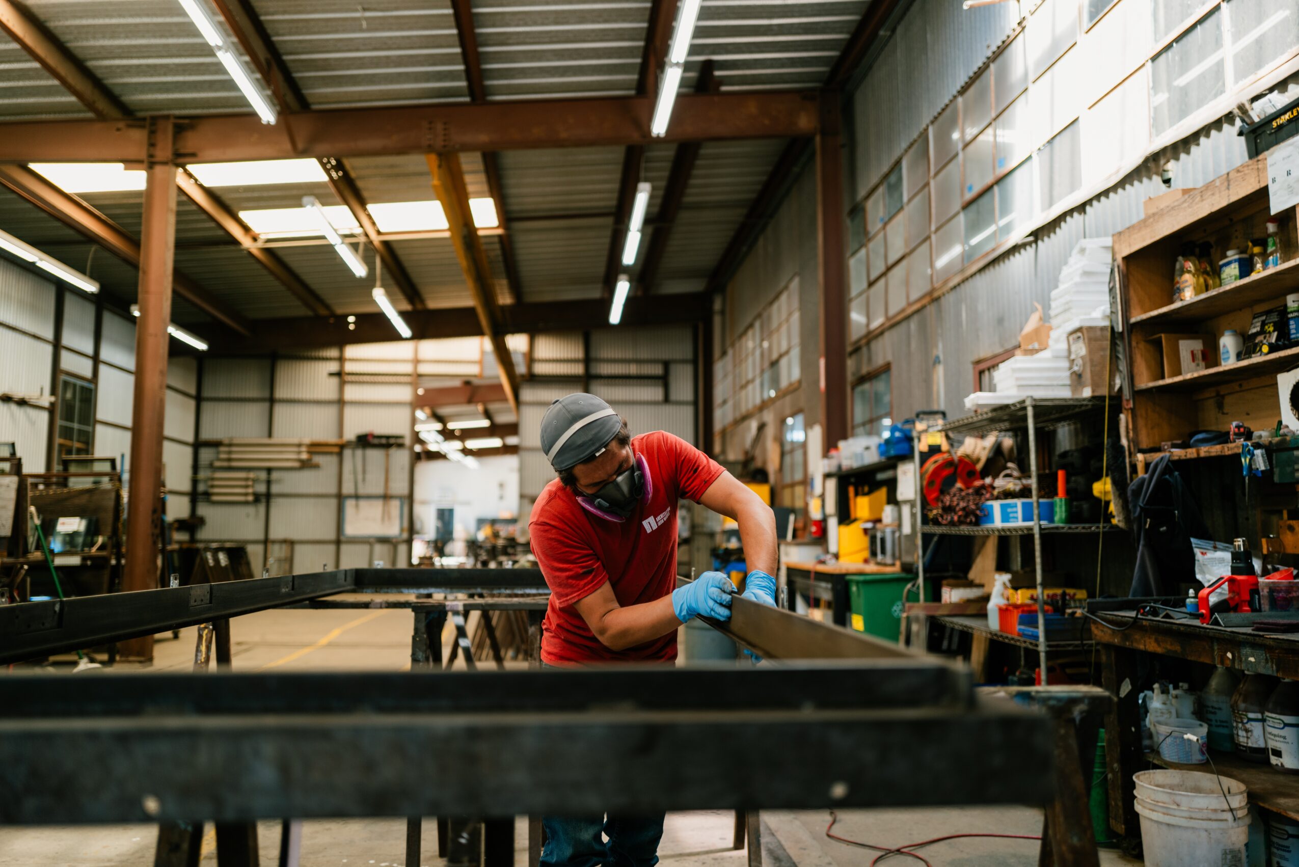 Person wearing a helmet and gloves working on a metal structure in a workshop with shelves and tools.