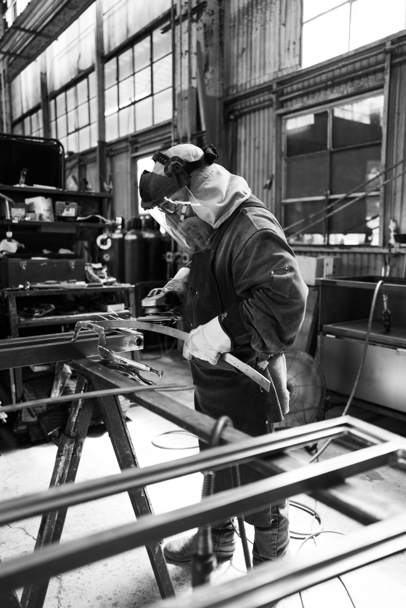 Two workers in a factory, one welding and the other assisting, with large windows and industrial equipment in the background.