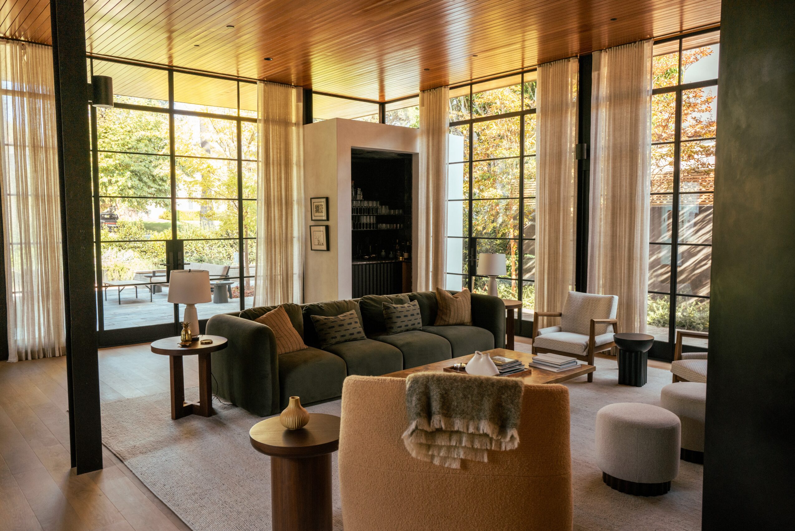 Living room with large windows, sofa, armchair, and side tables, illuminated by natural light, with a wooden ceiling and view outside.