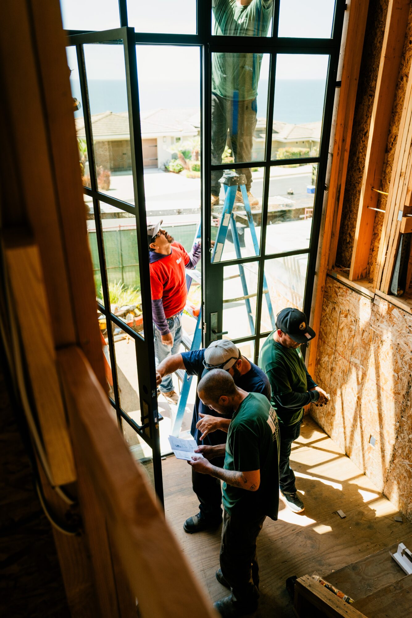 People working on a ladder outside a building viewed through a window, with sunlight streaming in. Word count: 20.