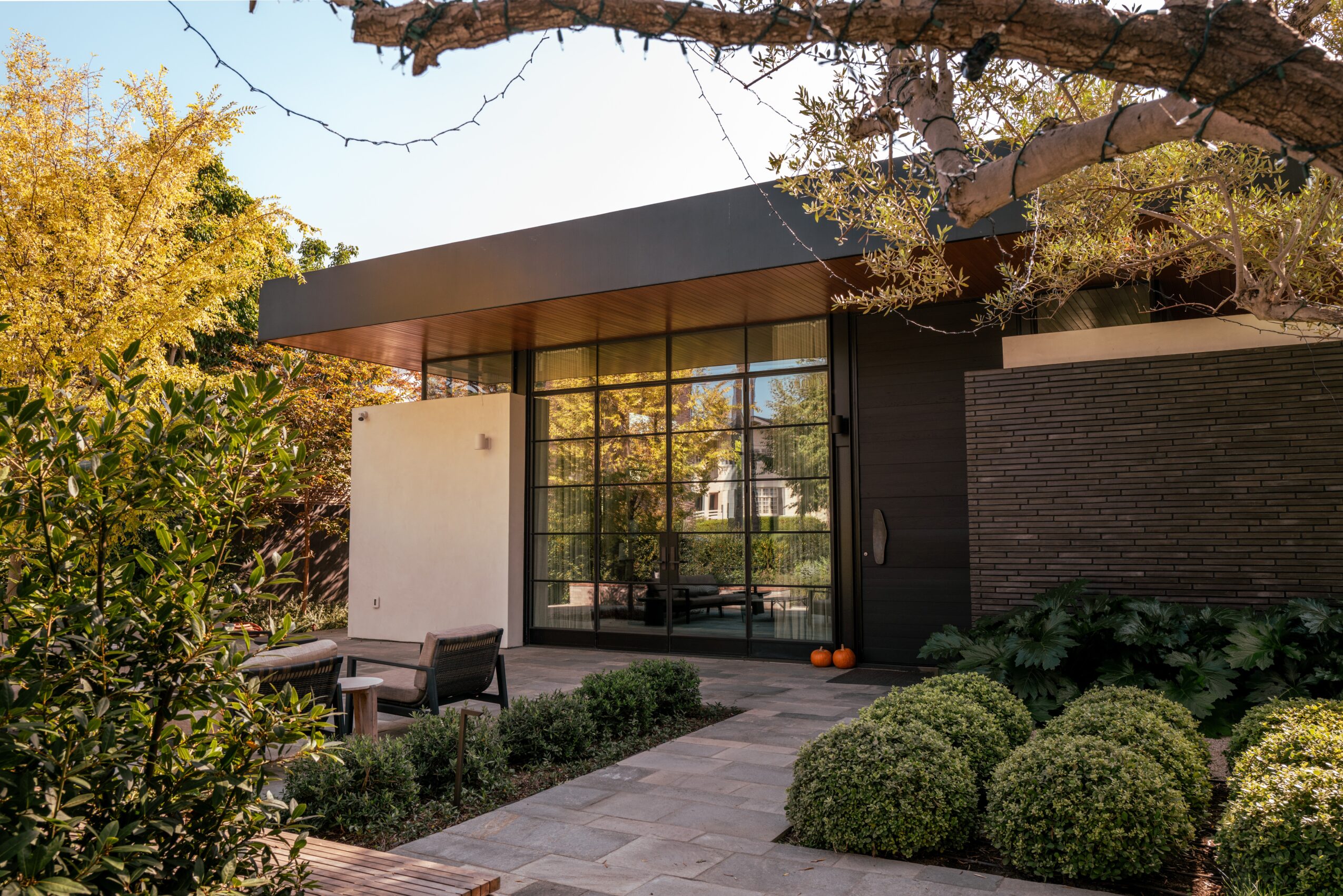 Modern house with large glass windows, surrounded by trees and bushes, with a paved walkway and outdoor seating area.