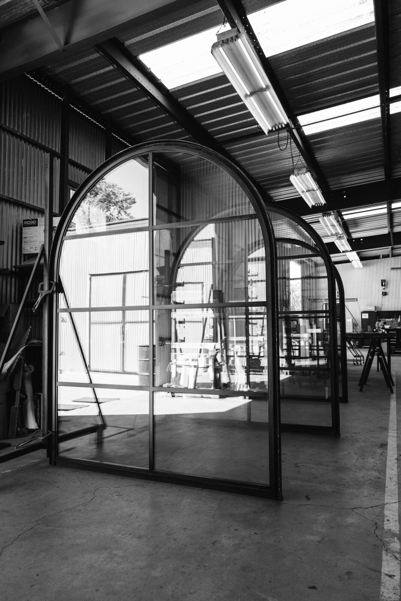 A black and white photo of a greenhouse structure inside a warehouse, with large windows and metal framing, and industrial lighting.