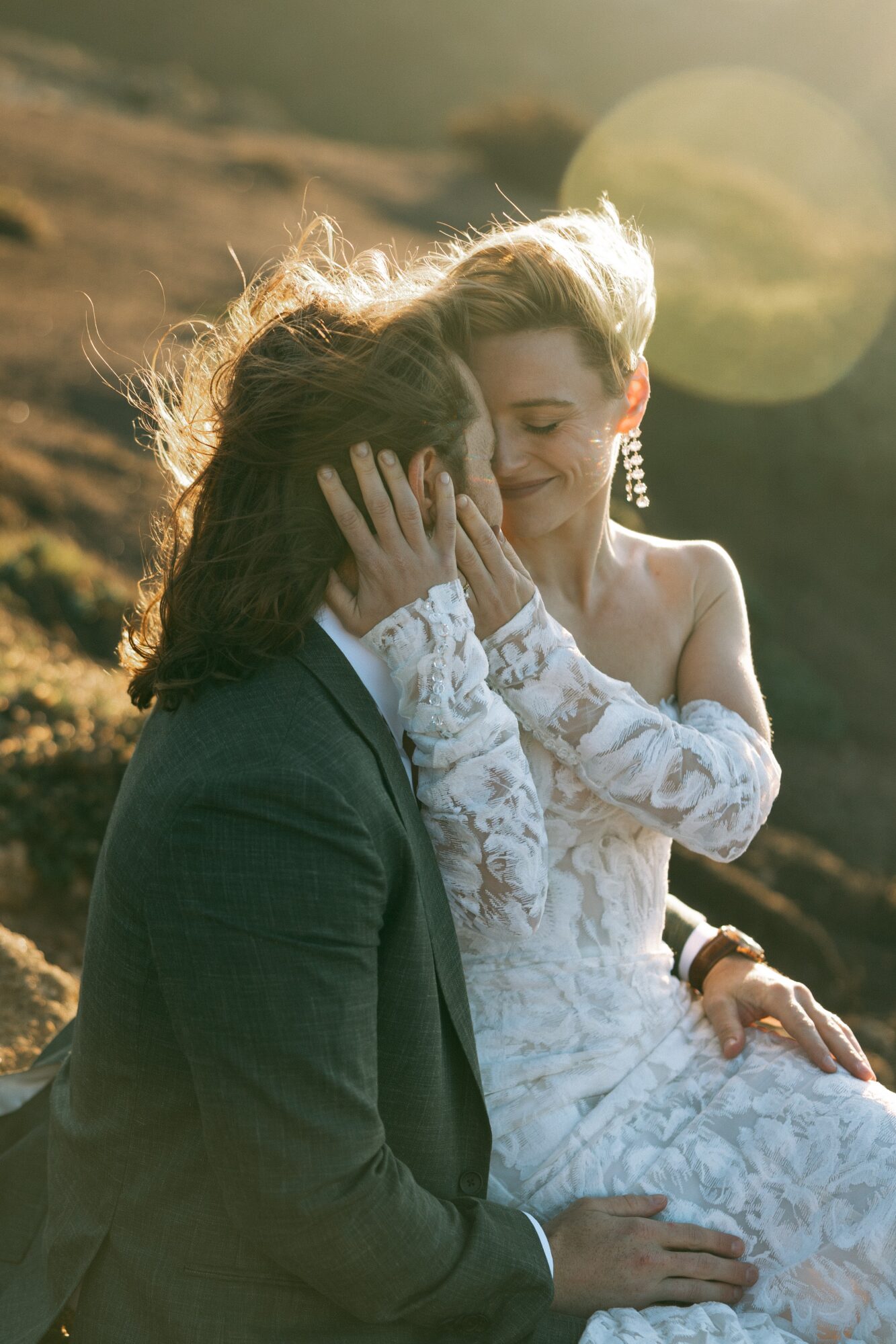 Two women in wedding attire share an intimate moment outdoors, with one touching the other's face and smiling.