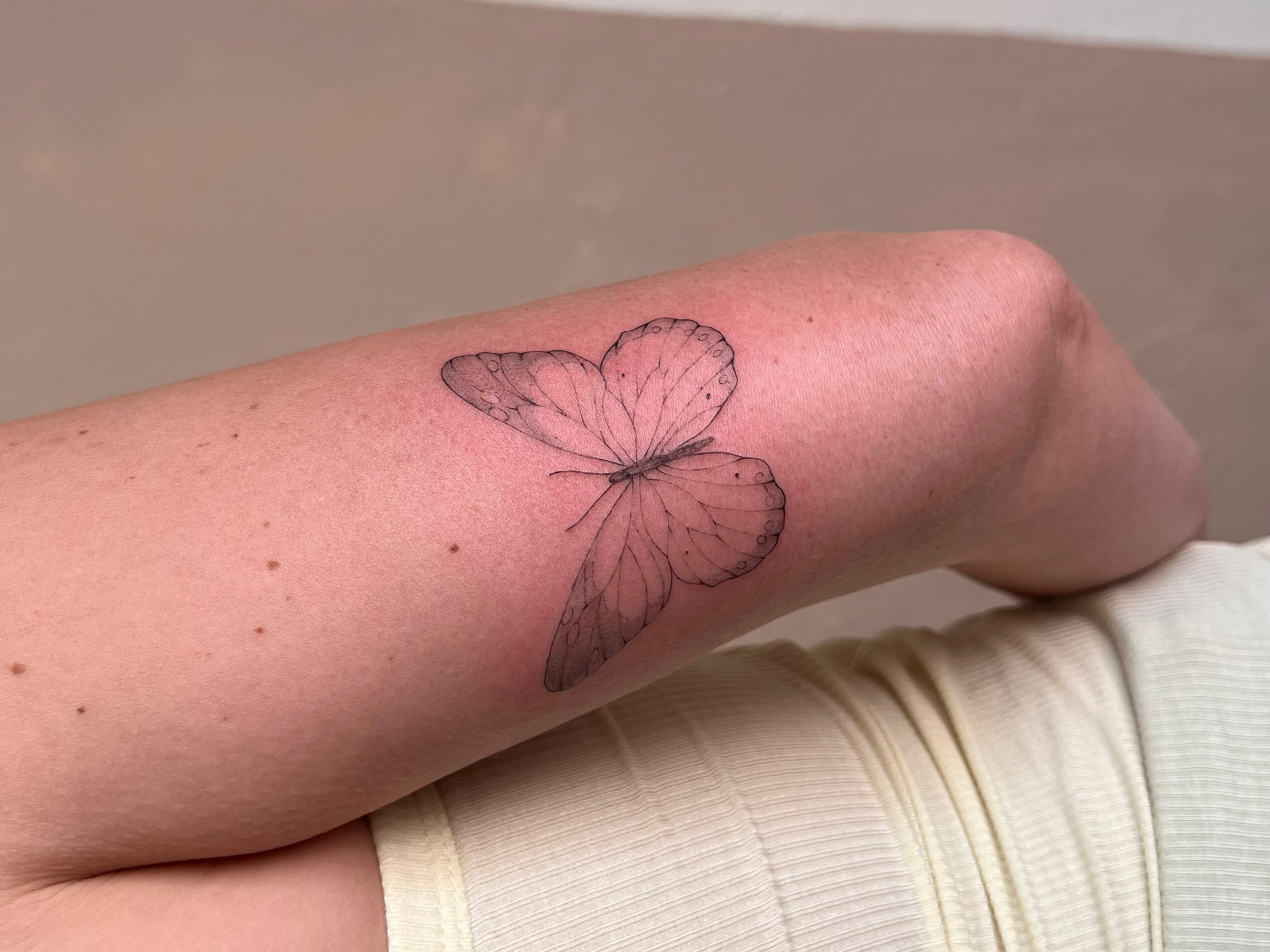 Forearm with a butterfly tattoo, skin appears smooth, background is plain, person wears a light-colored shirt.