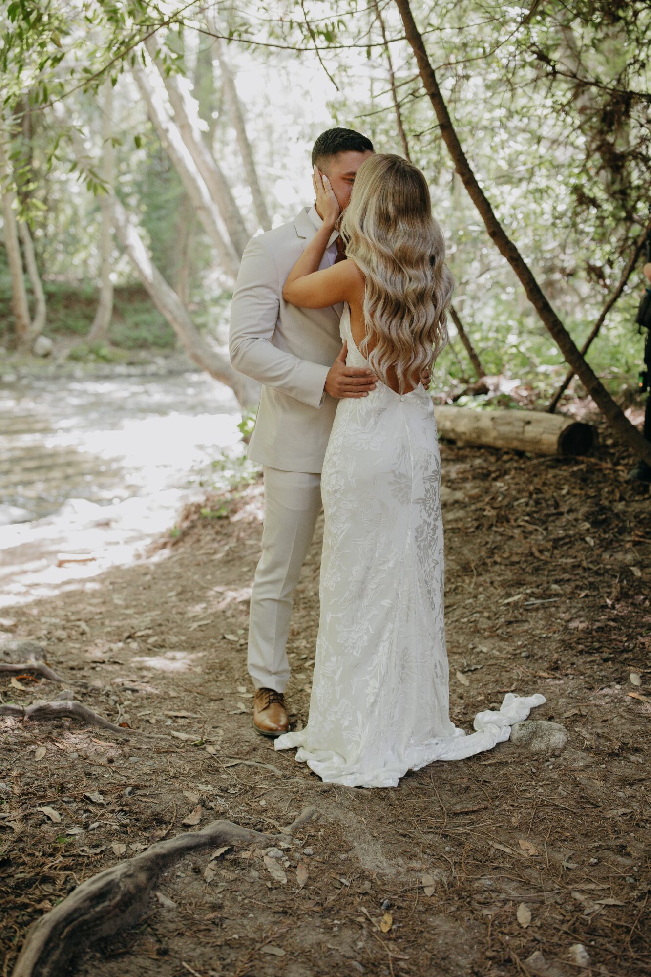 A couple in wedding attire embracing in a forested area with sunlight filtering through trees.