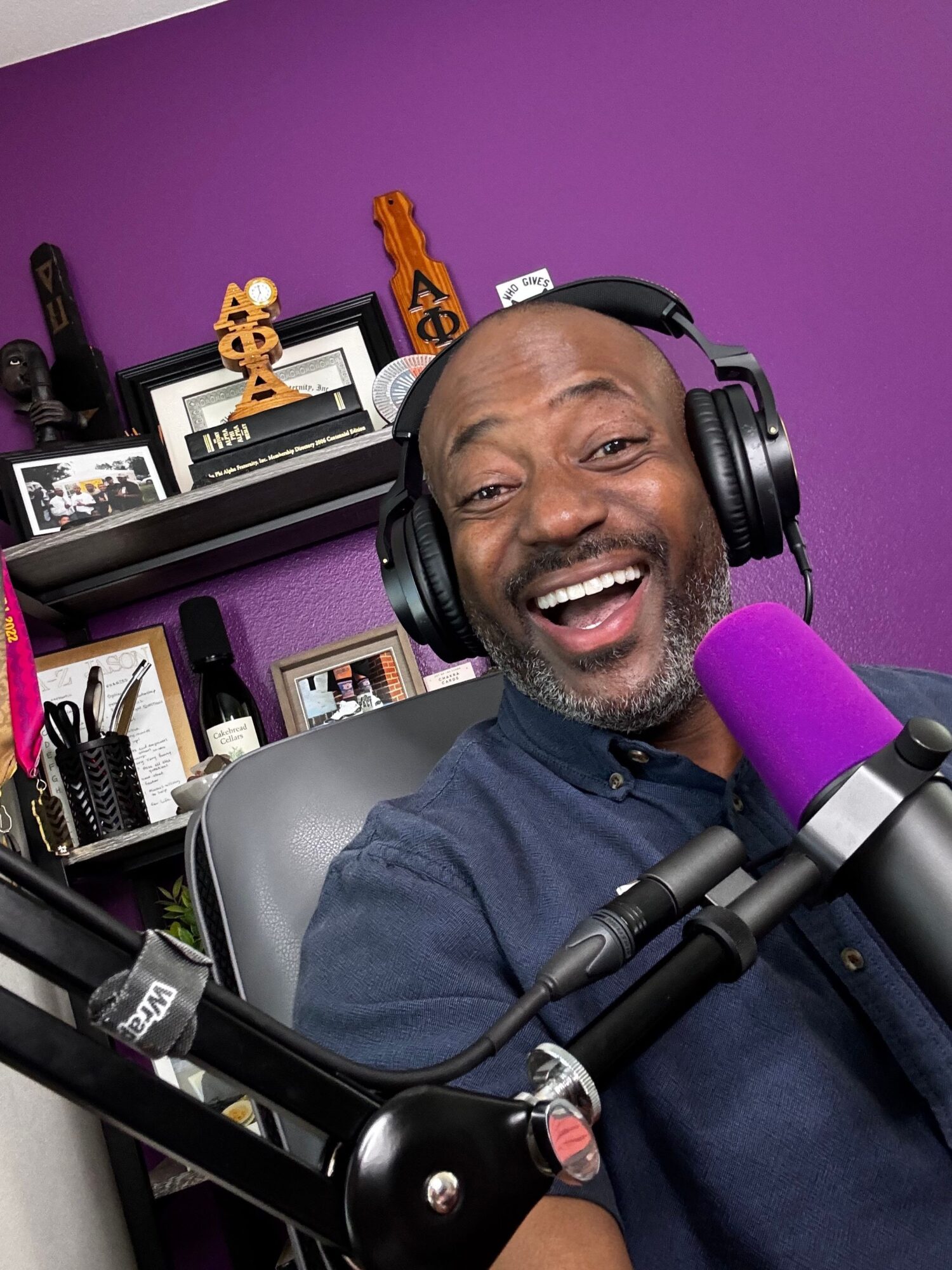 Man smiling with headphones speaking into a microphone in a purple room with shelves and decorations.