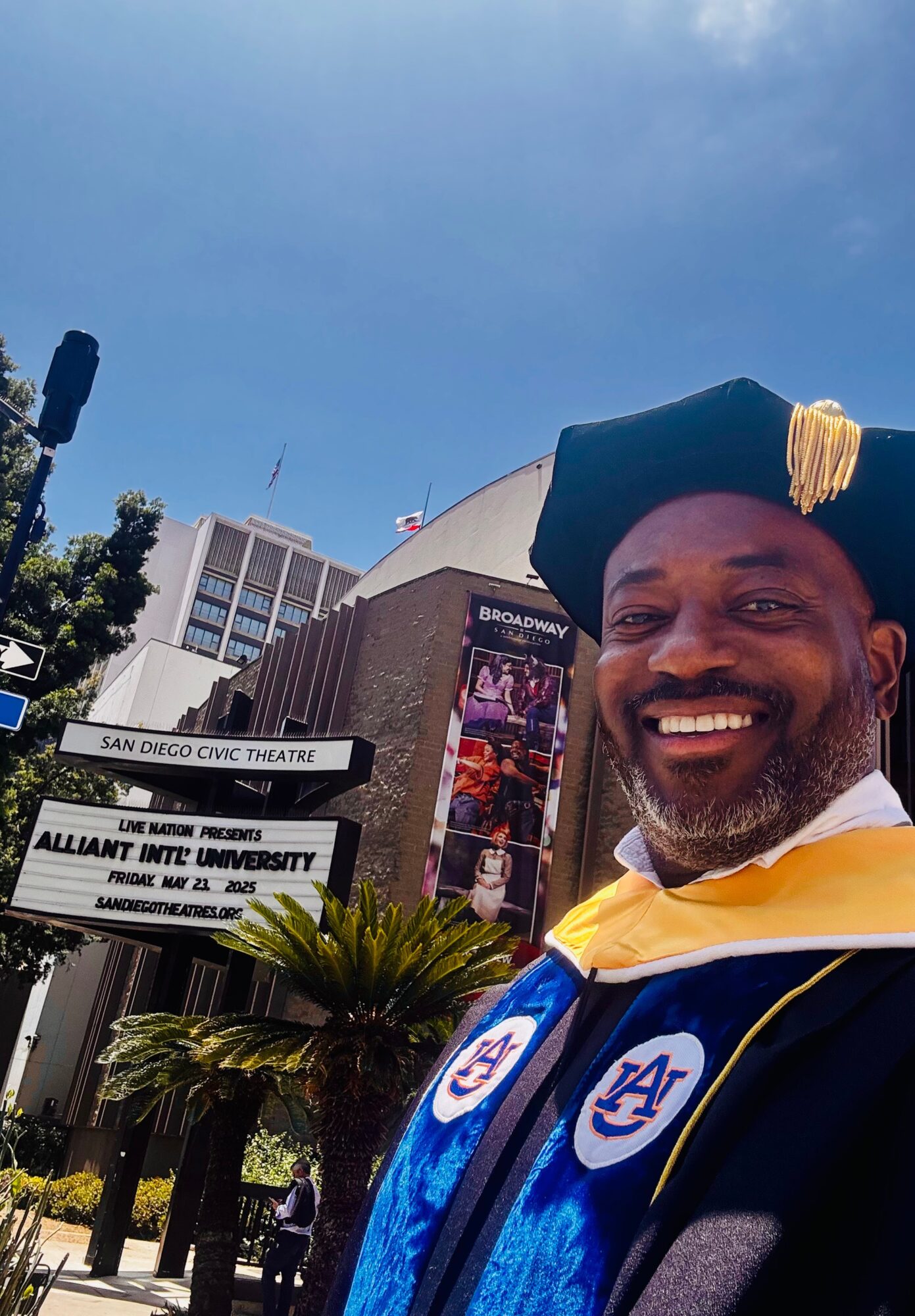 Smiling man in academic regalia outdoors with buildings and signs in the background.