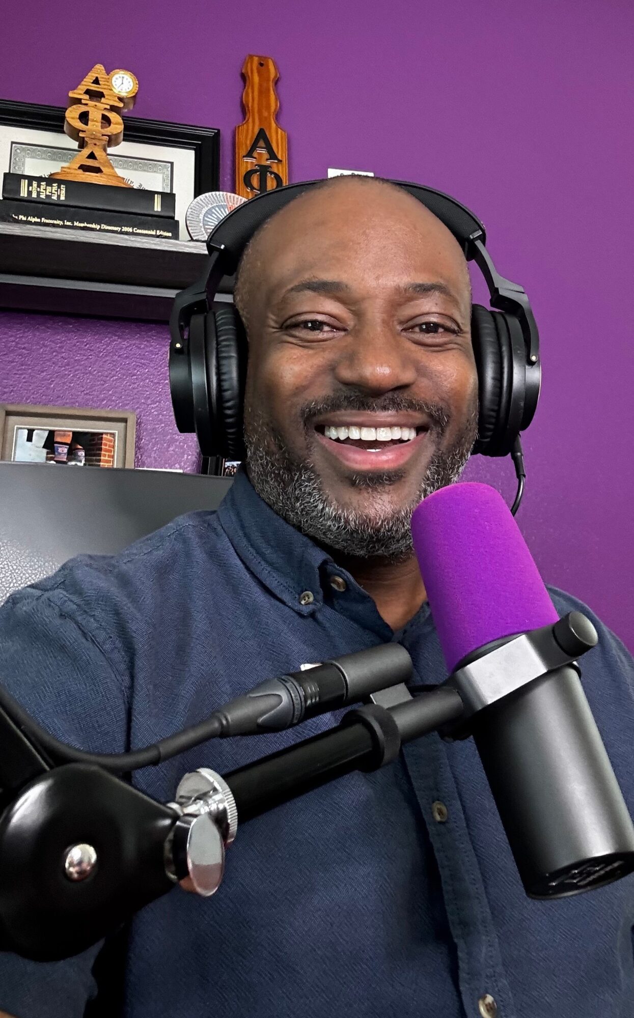 Smiling man wearing headphones in front of purple background with microphone, bookshelf, and decorative items behind him.