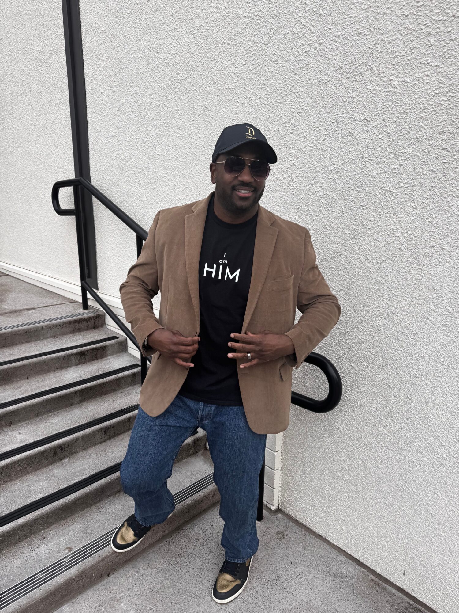 Person standing on stairs, wearing a beige blazer, black shirt, jeans, and sneakers, holding a phone, with a white wall background.