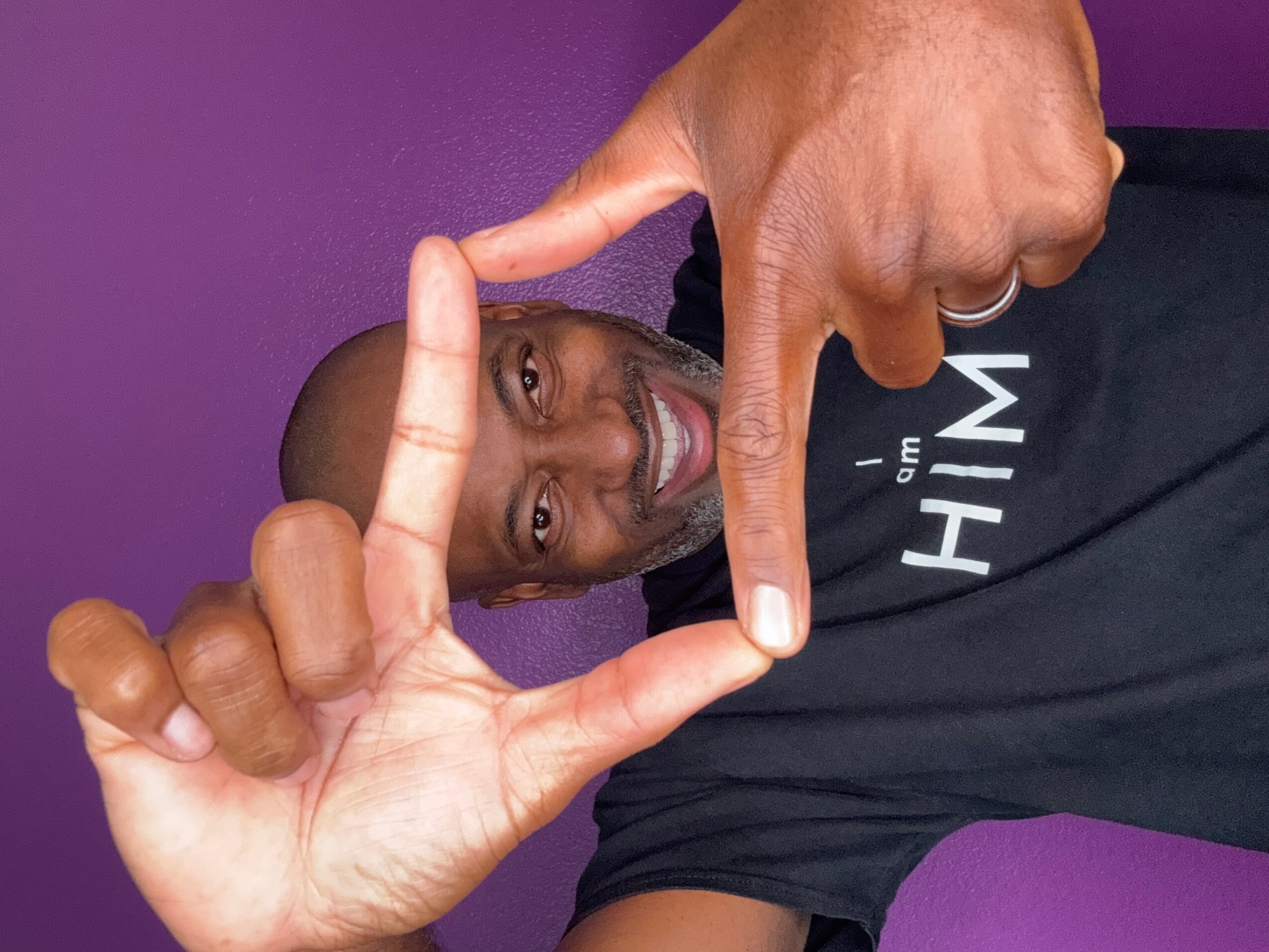 Person smiling, making a frame with hands, purple background, wearing a black shirt with white text, head tilted, looking through the frame.
