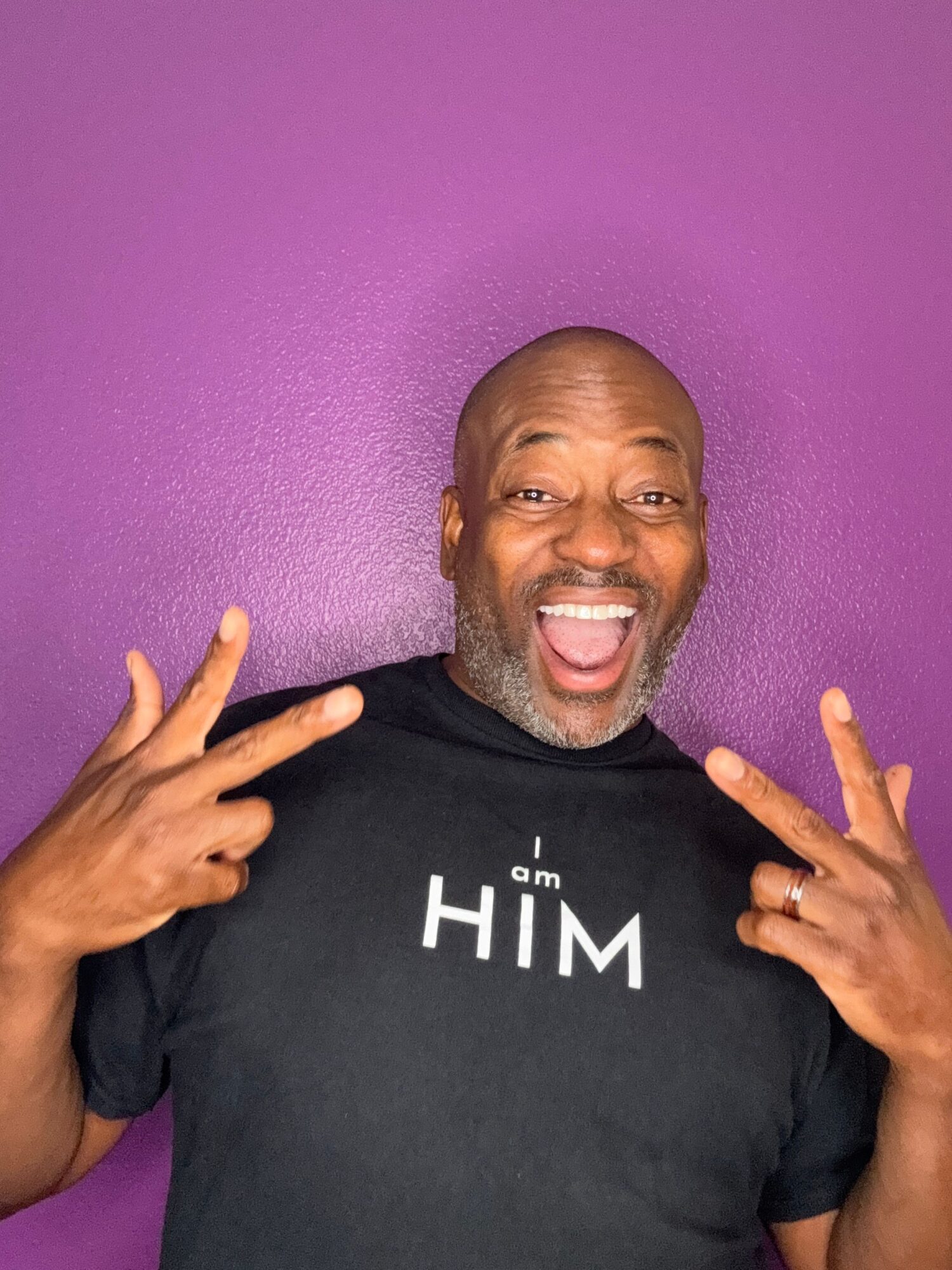 Man smiling and making hand gestures against a purple wall, wearing a black shirt with white text.
