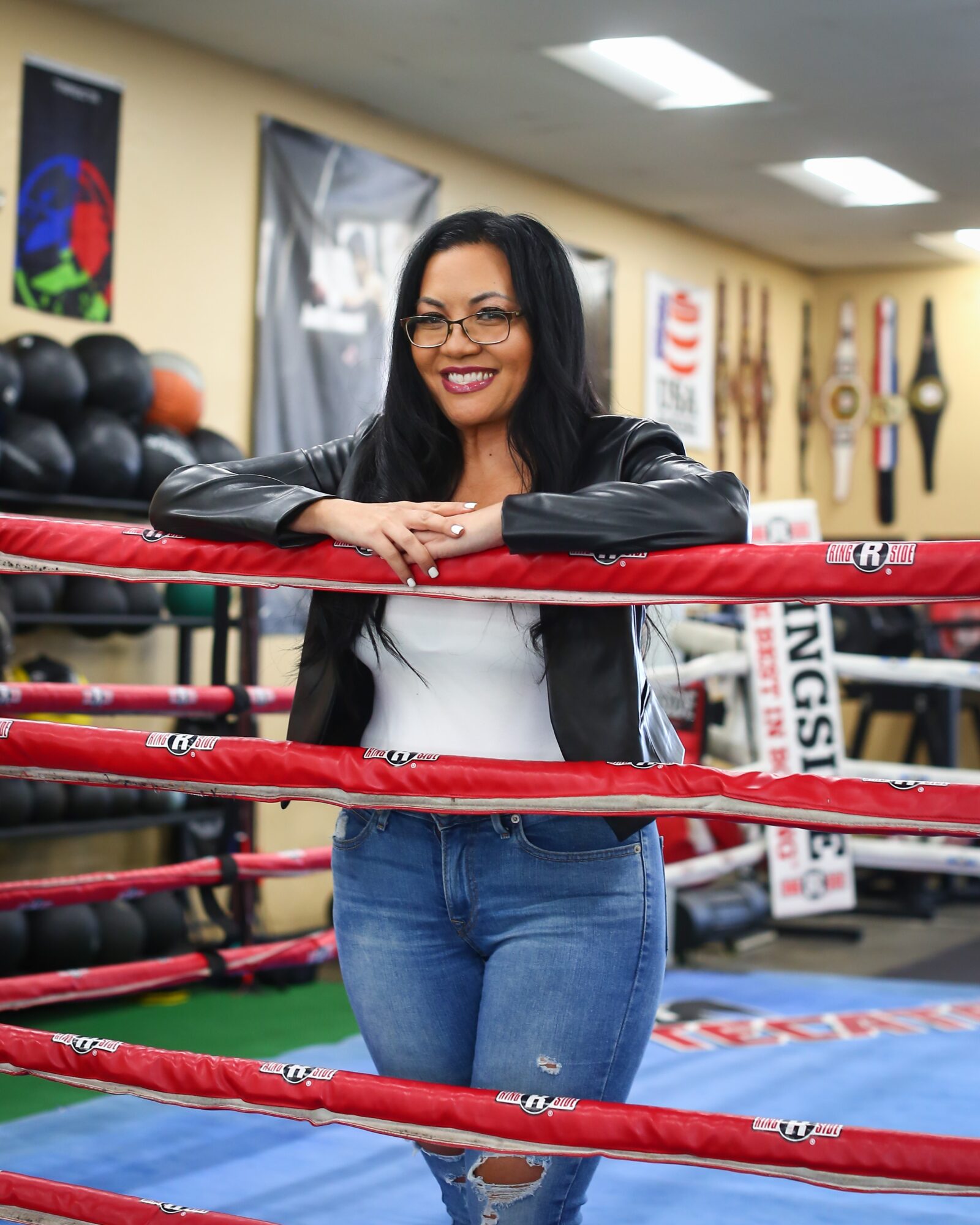 Smiling woman with glasses leaning on boxing ring ropes in gym, surrounded by fitness equipment and posters.