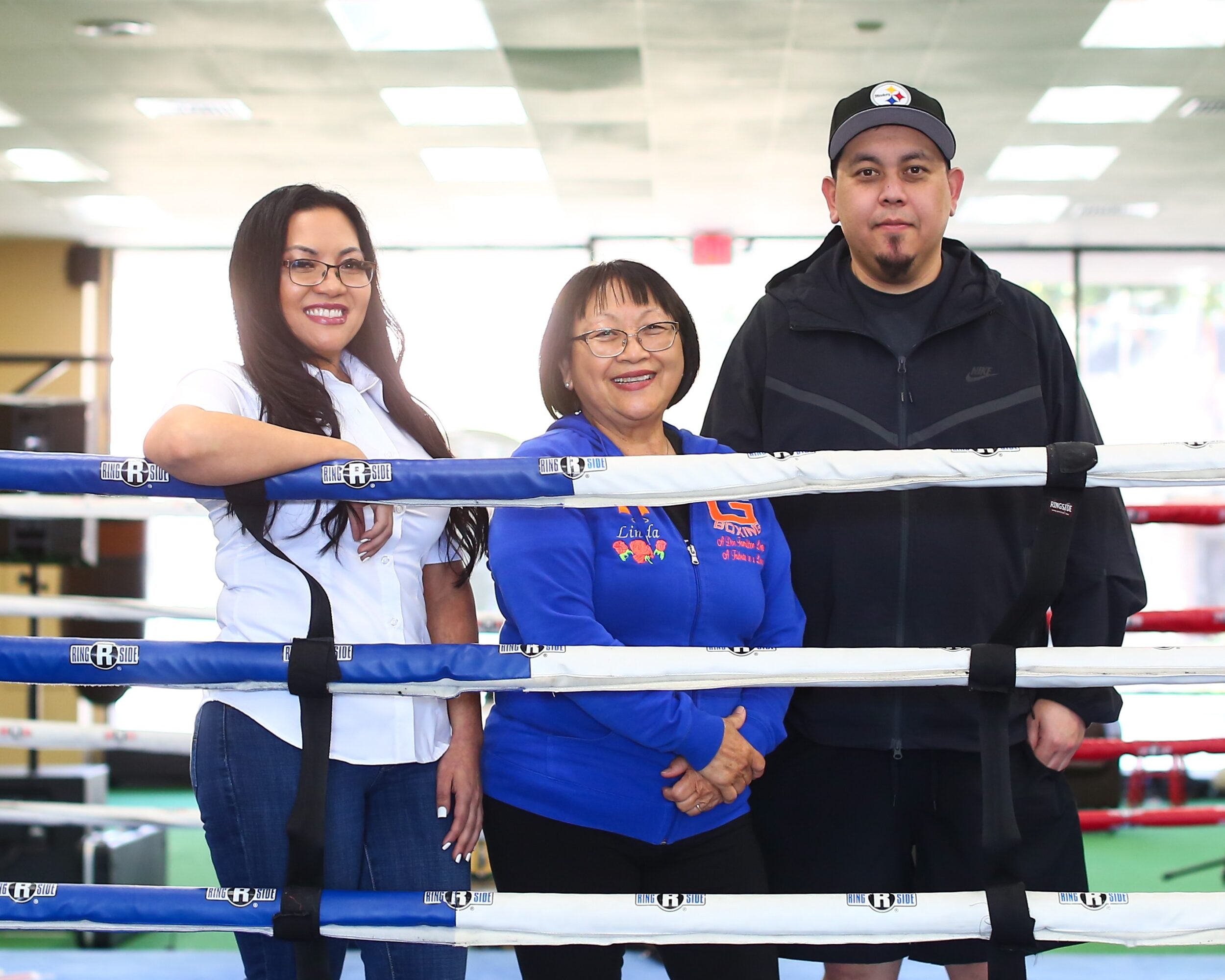 Three people standing behind boxing ring ropes in a gym, smiling, with ceiling lights overhead.