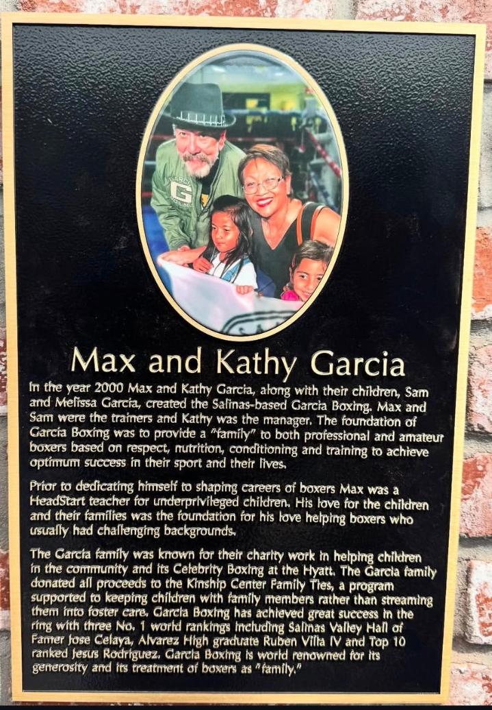 Photograph of three people, two adults and a child, smiling and posing together, with a black background and an oval frame. The image is part of a memorial plaque with text below.