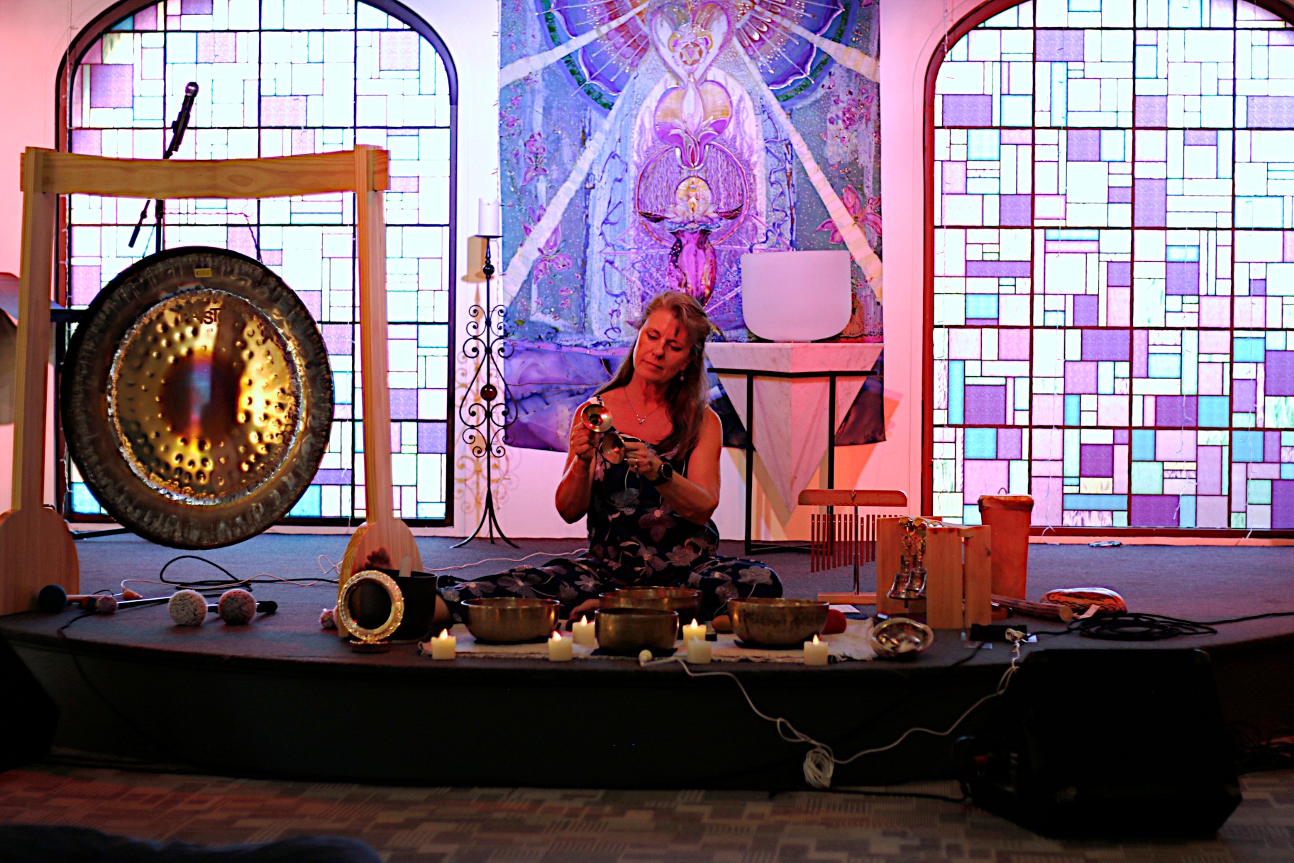 Person sitting cross-legged with musical instruments and objects on a stage, colorful stained glass and artwork behind, illuminated by vibrant lights.