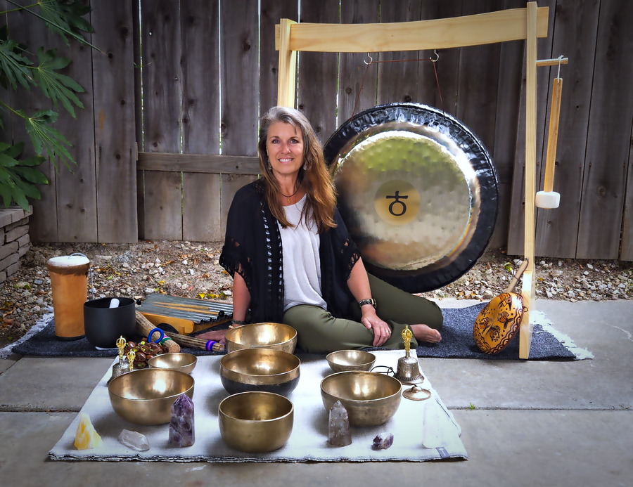 Woman sitting on ground with singing bowls and gong outdoors, wooden fence background, plants, and a mat.