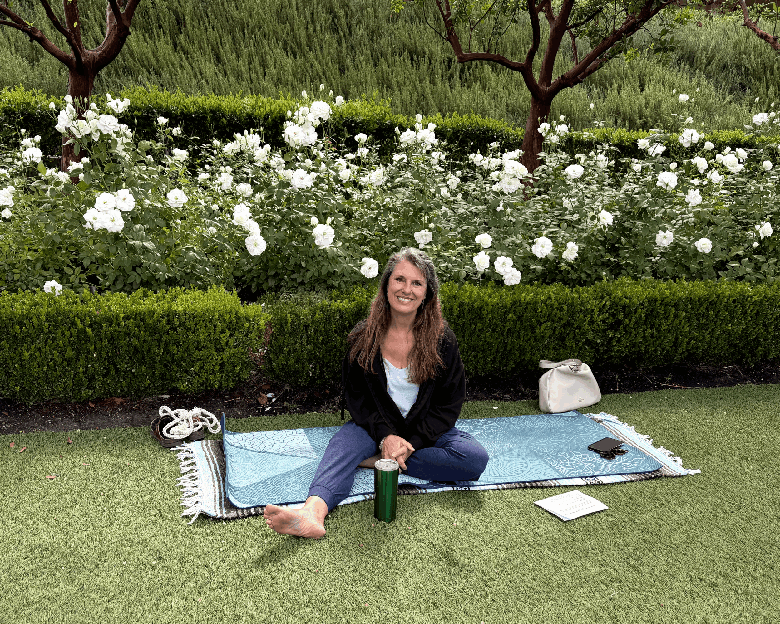 Young woman sitting on a mat outdoors with flowers and trees in the background, smiling, with a water bottle and a book nearby.