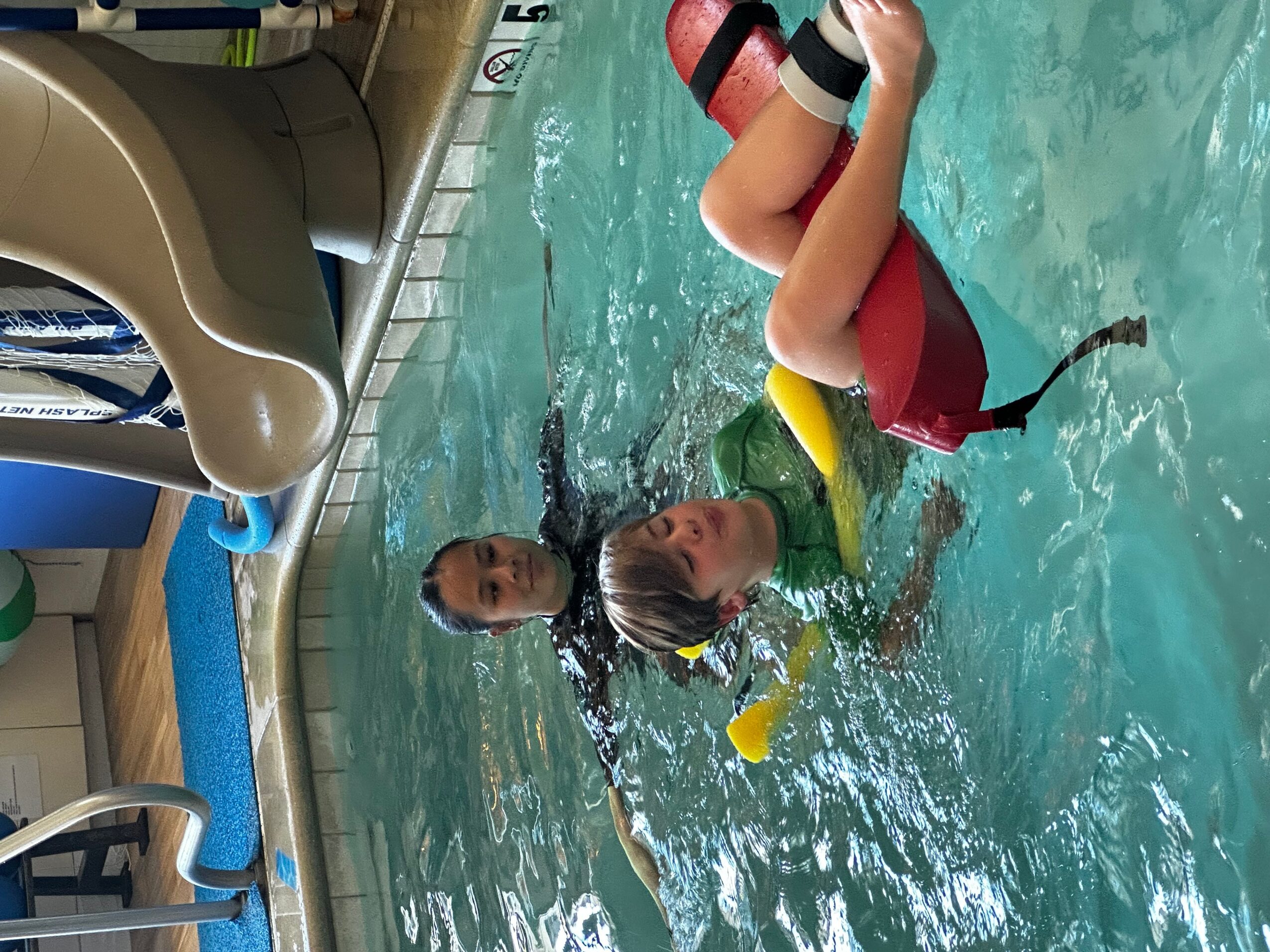 Two children swimming in a pool, one with a yellow float, the other with a red float, near pool edge.