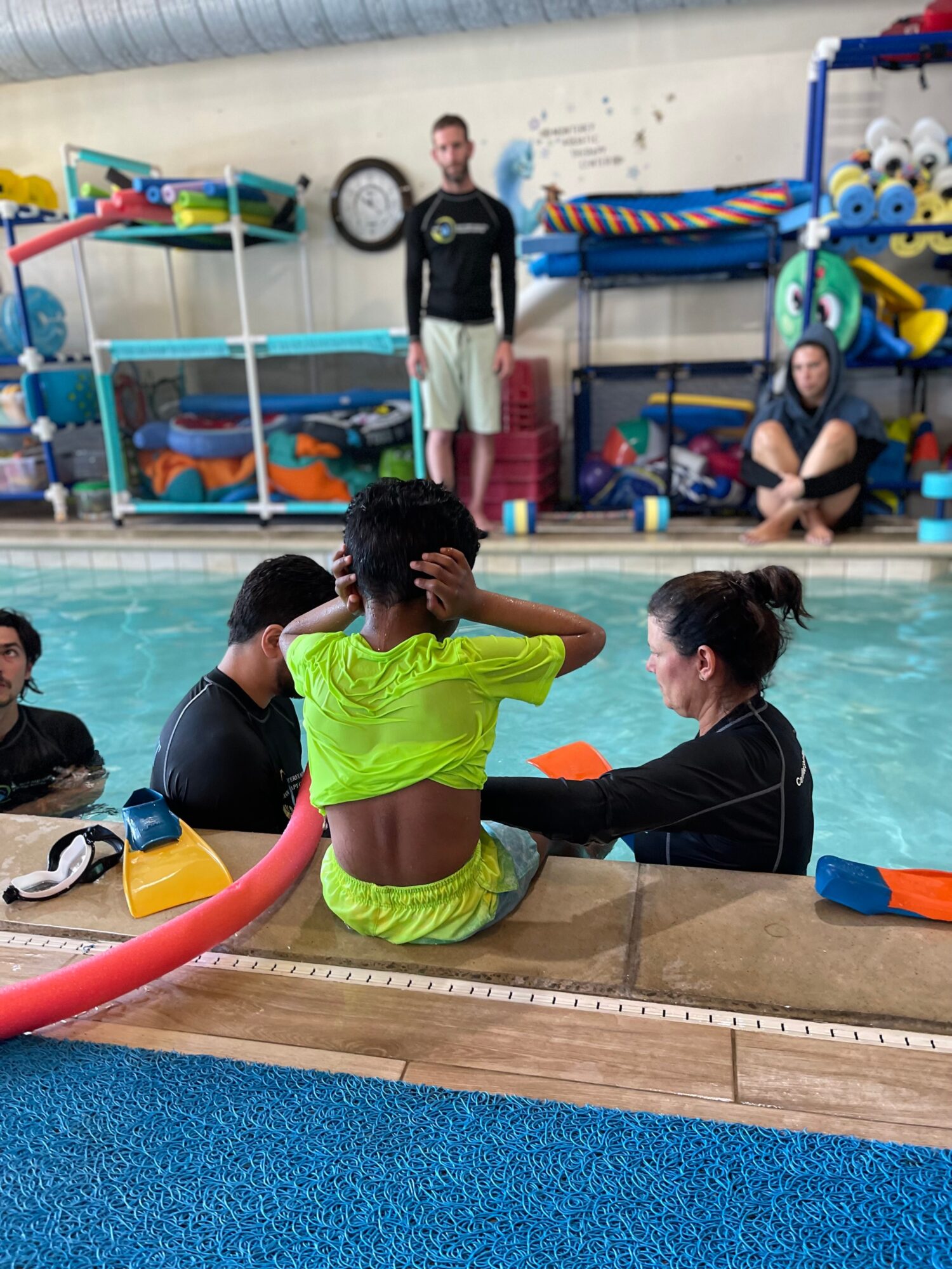 Person in bright green clothing sitting on poolside, with others in the pool, and a person standing in the background near pool equipment.