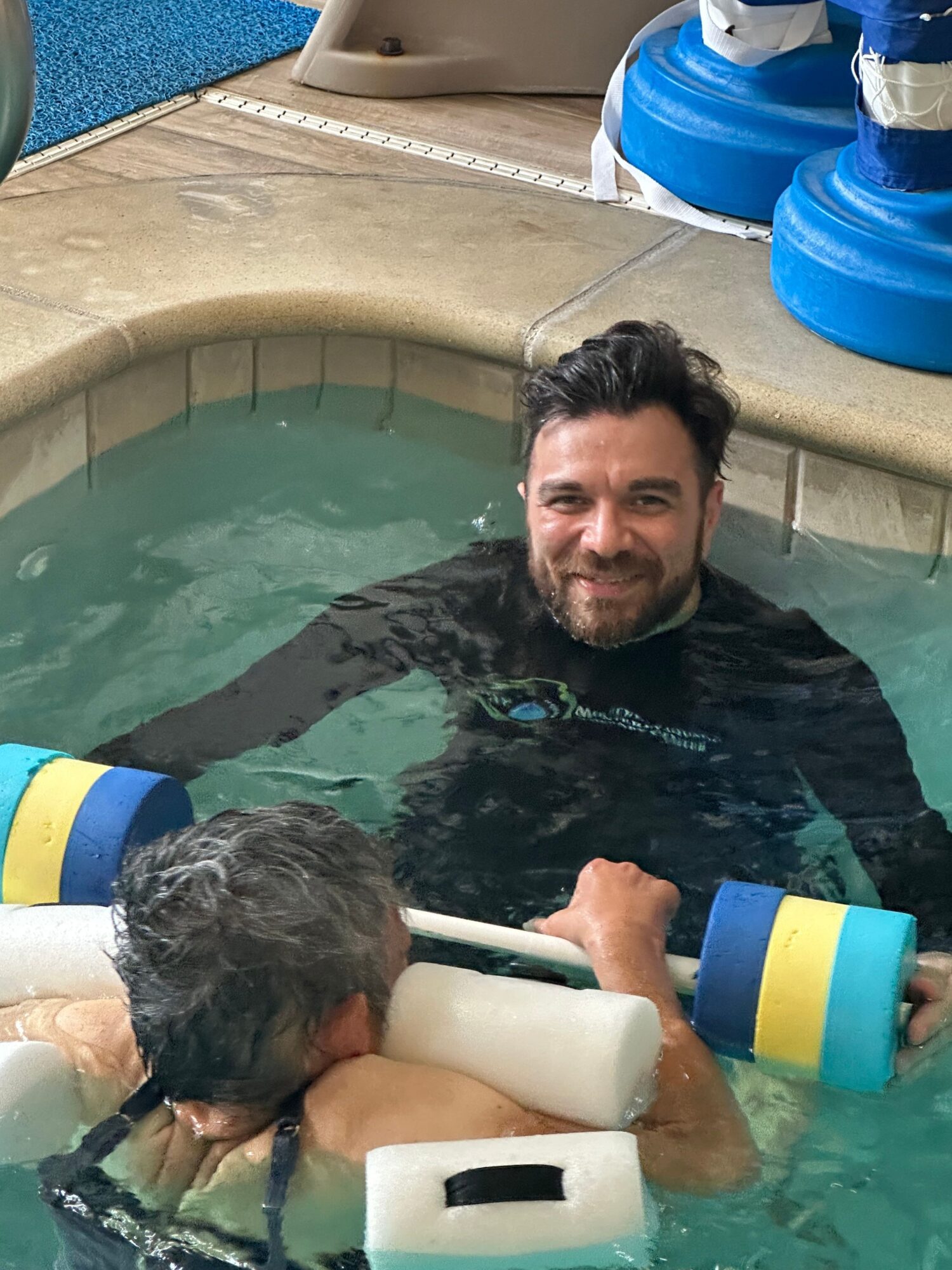 Man in a pool with a child, both holding foam pool noodles, surrounded by pool equipment, smiling and enjoying water.
