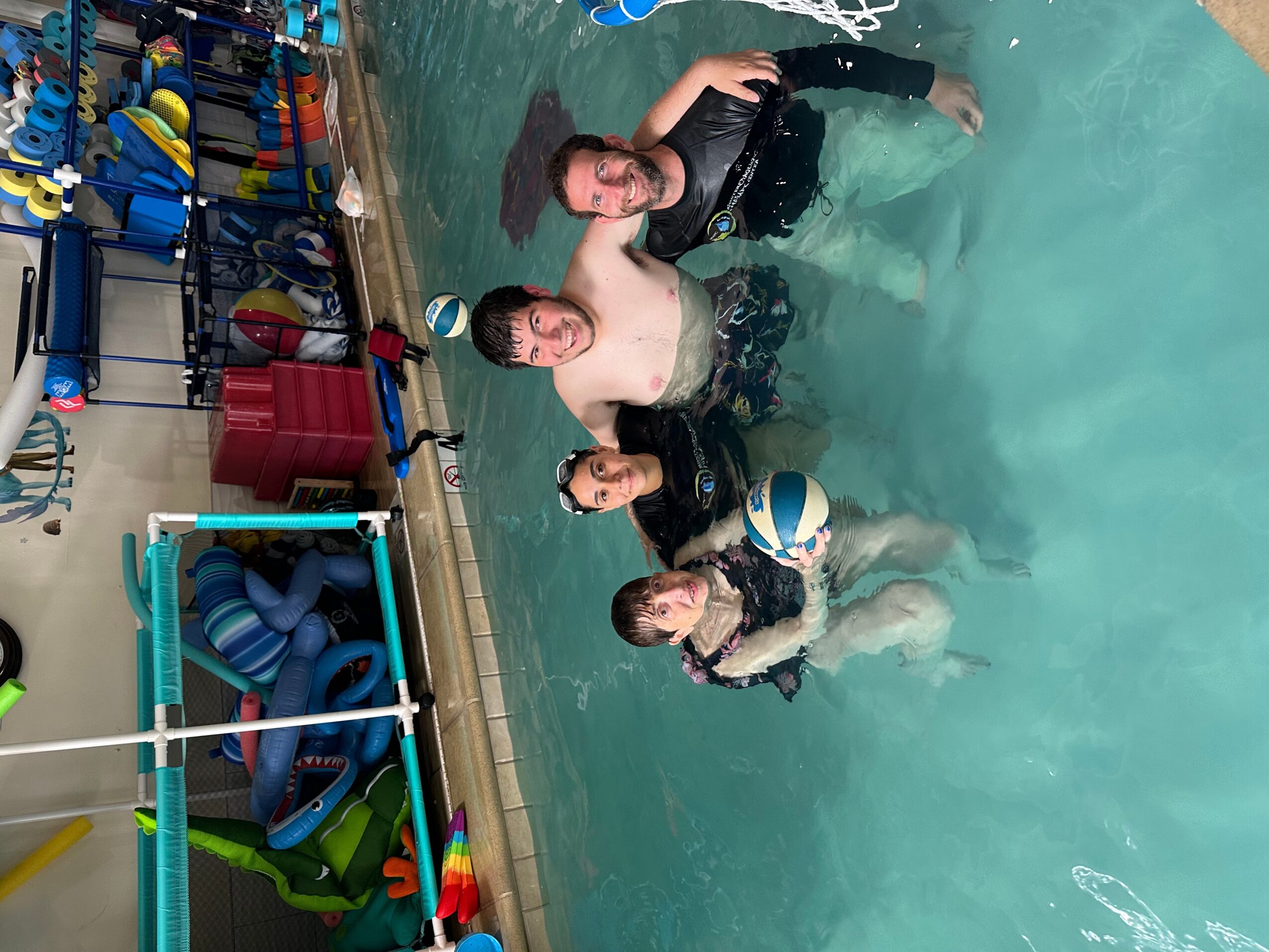 Four people in a swimming pool, holding a ball, with pool toys and equipment on the side.