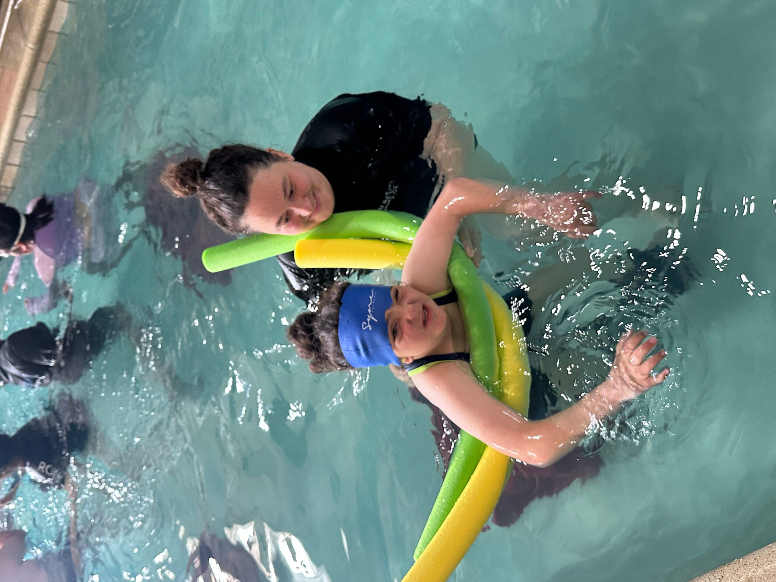Two children in a swimming pool, one with a blue swim cap, using a yellow and green floatation device, smiling and playing.