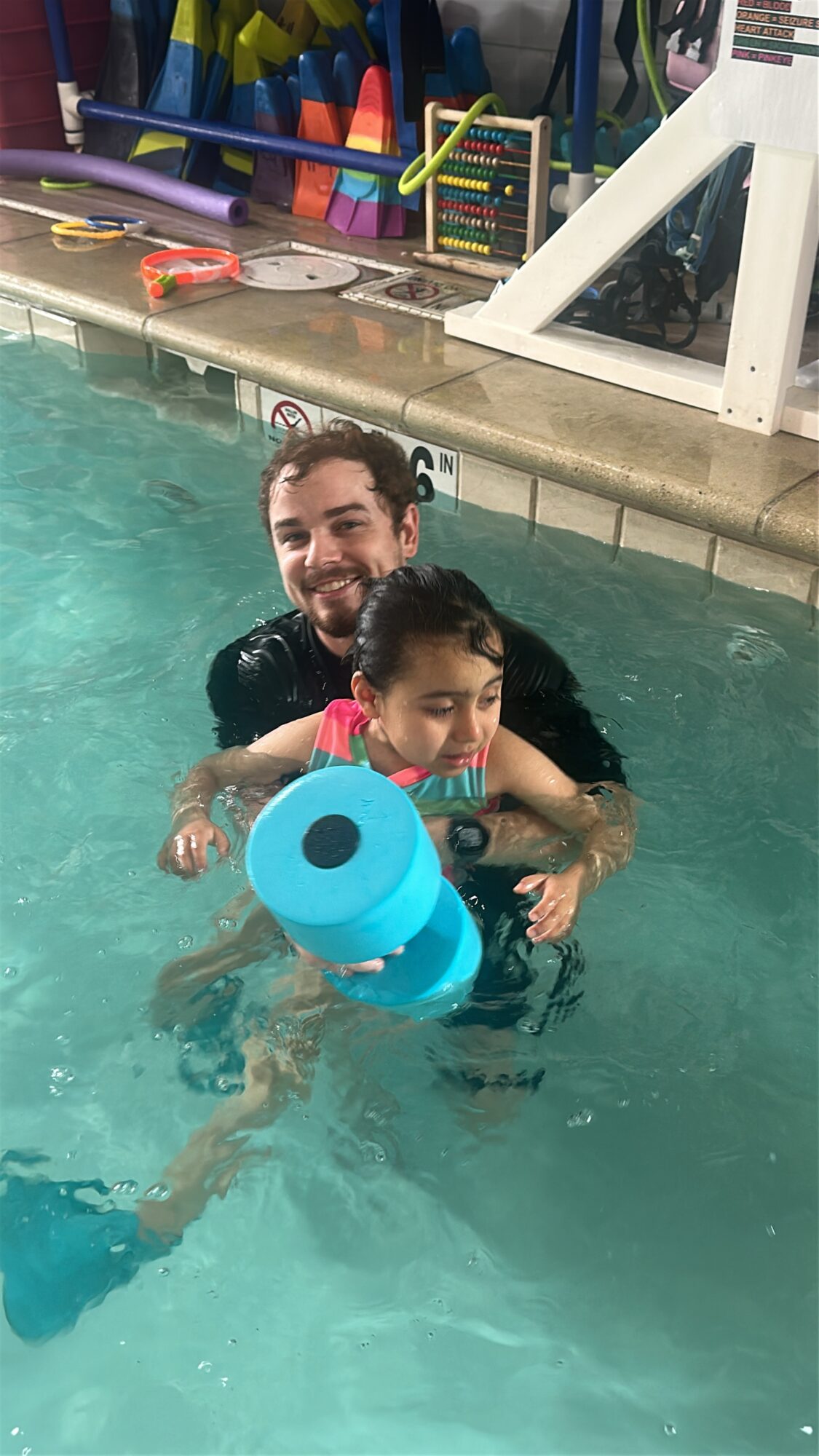 Adult and child in swimming pool holding a blue pool float, smiling, with pool equipment in background.