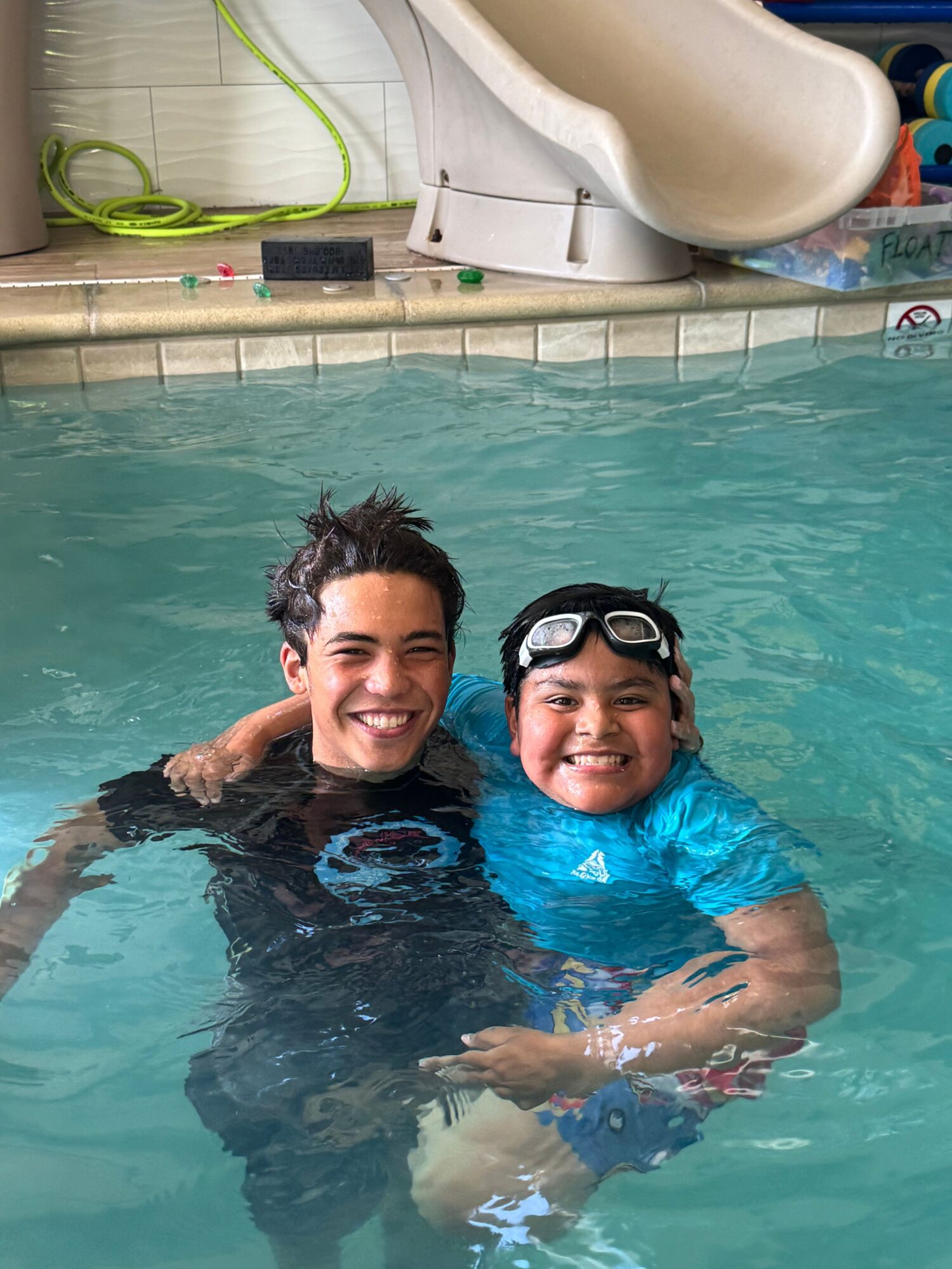Two people in a swimming pool, smiling, with one wearing goggles, close together, water around them.