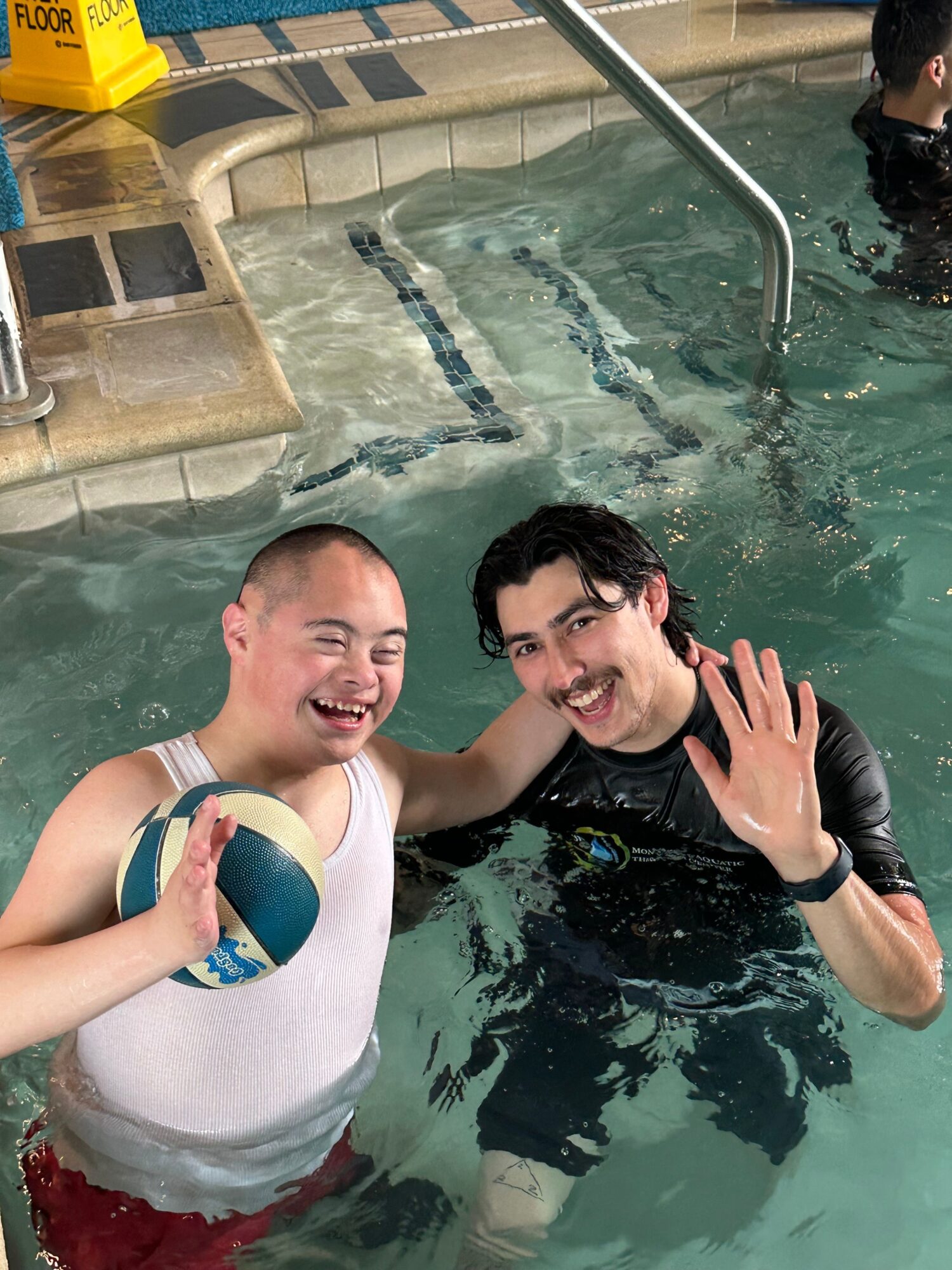 Two people in a swimming pool, smiling and holding a volleyball, with one person making a peace sign.