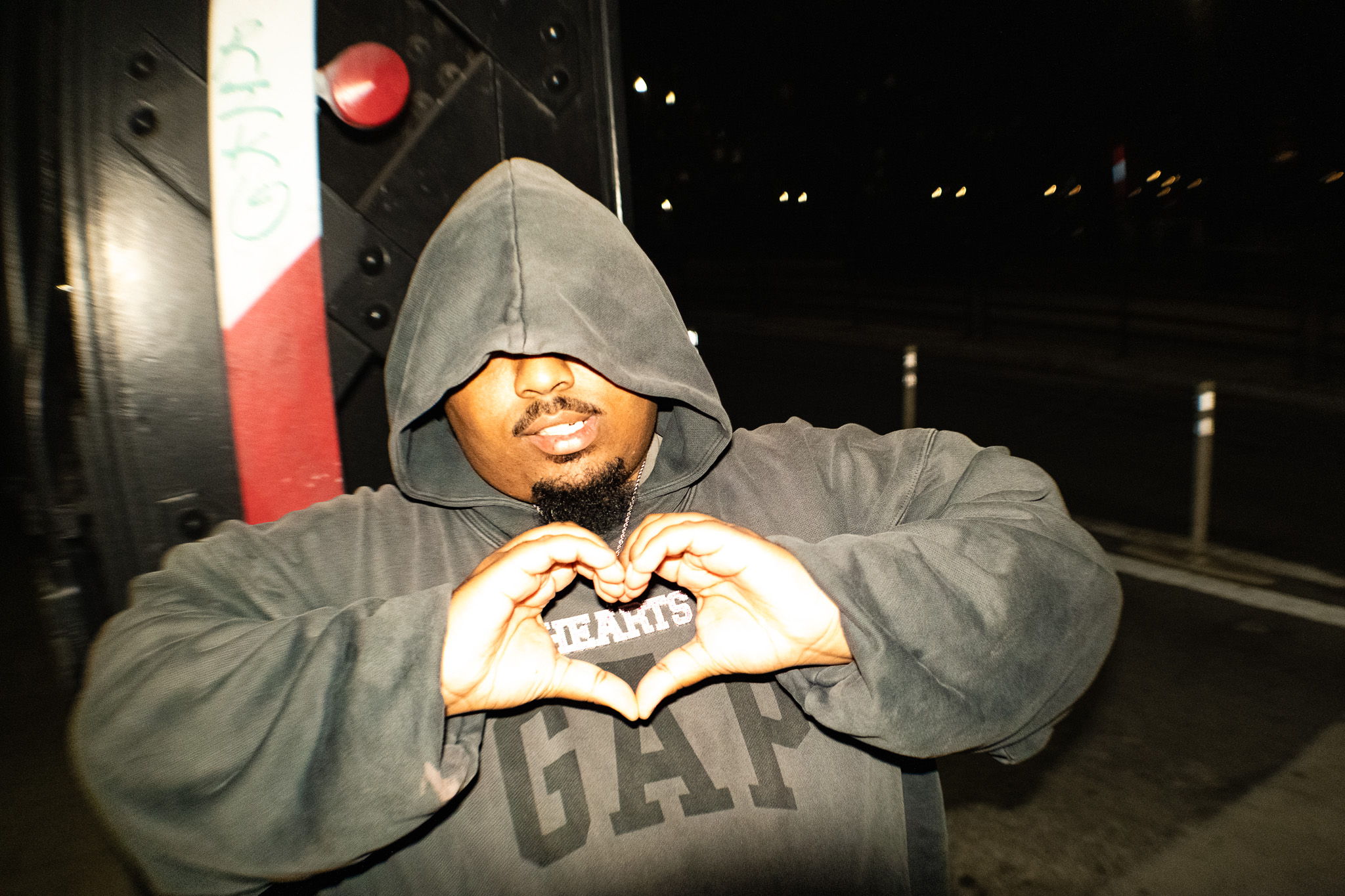 Person wearing a hoodie making a heart shape with hands at night outdoors.