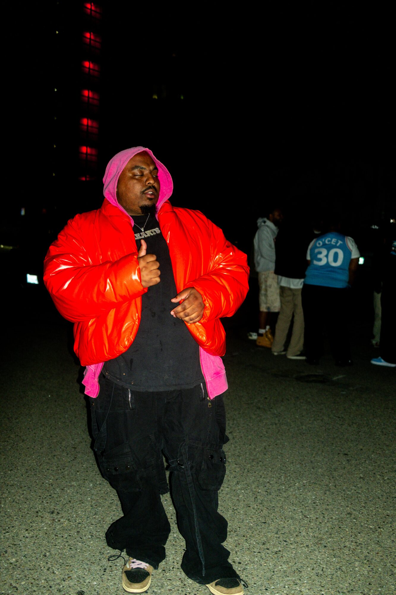 Person wearing a red jacket and pink hood holding a microphone at night, with people in the background.