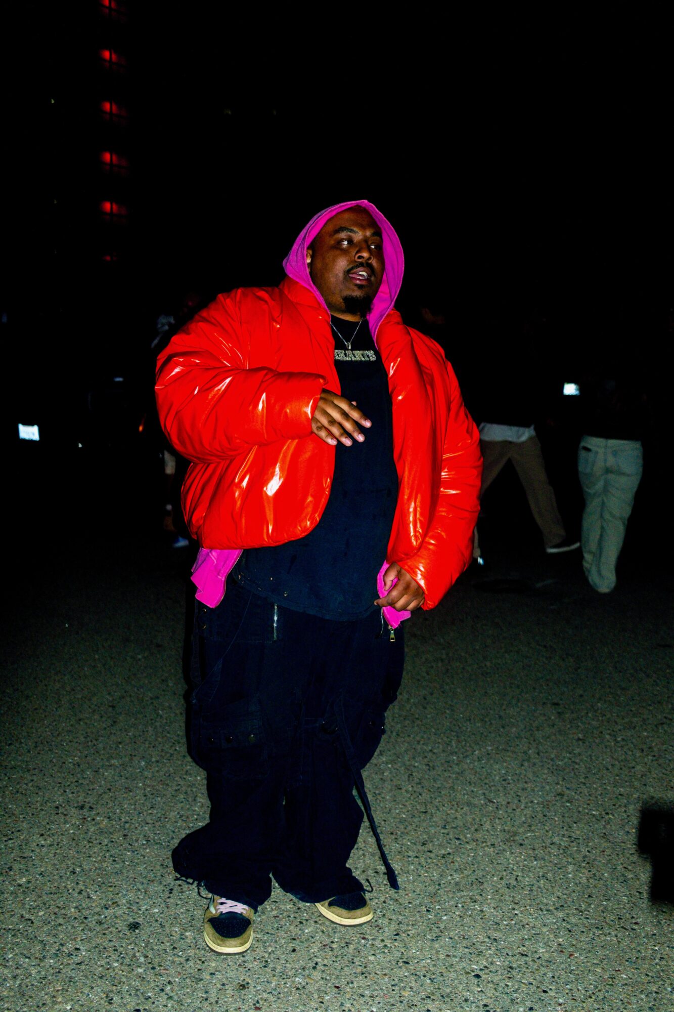 Person wearing a red puffy jacket, pink hoodie, and dark pants standing outdoors at night, with other people in the background.