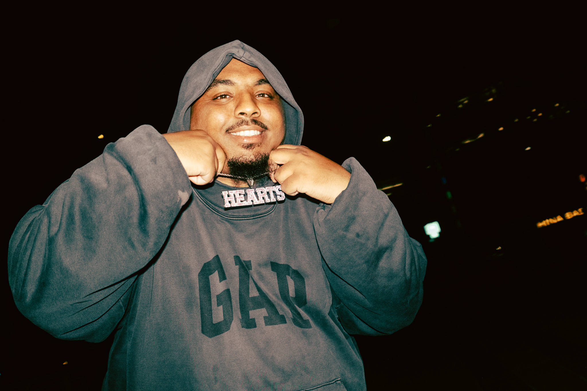 Person wearing a hoodie with hands near face, smiling, dark background, city lights visible.