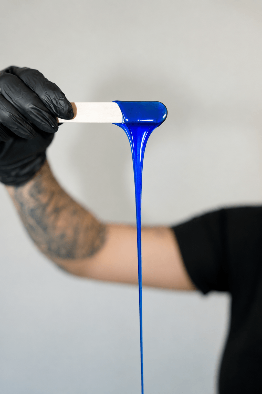 Person holding a test tube with blue liquid dripping, arm tattoo visible, background plain gray.