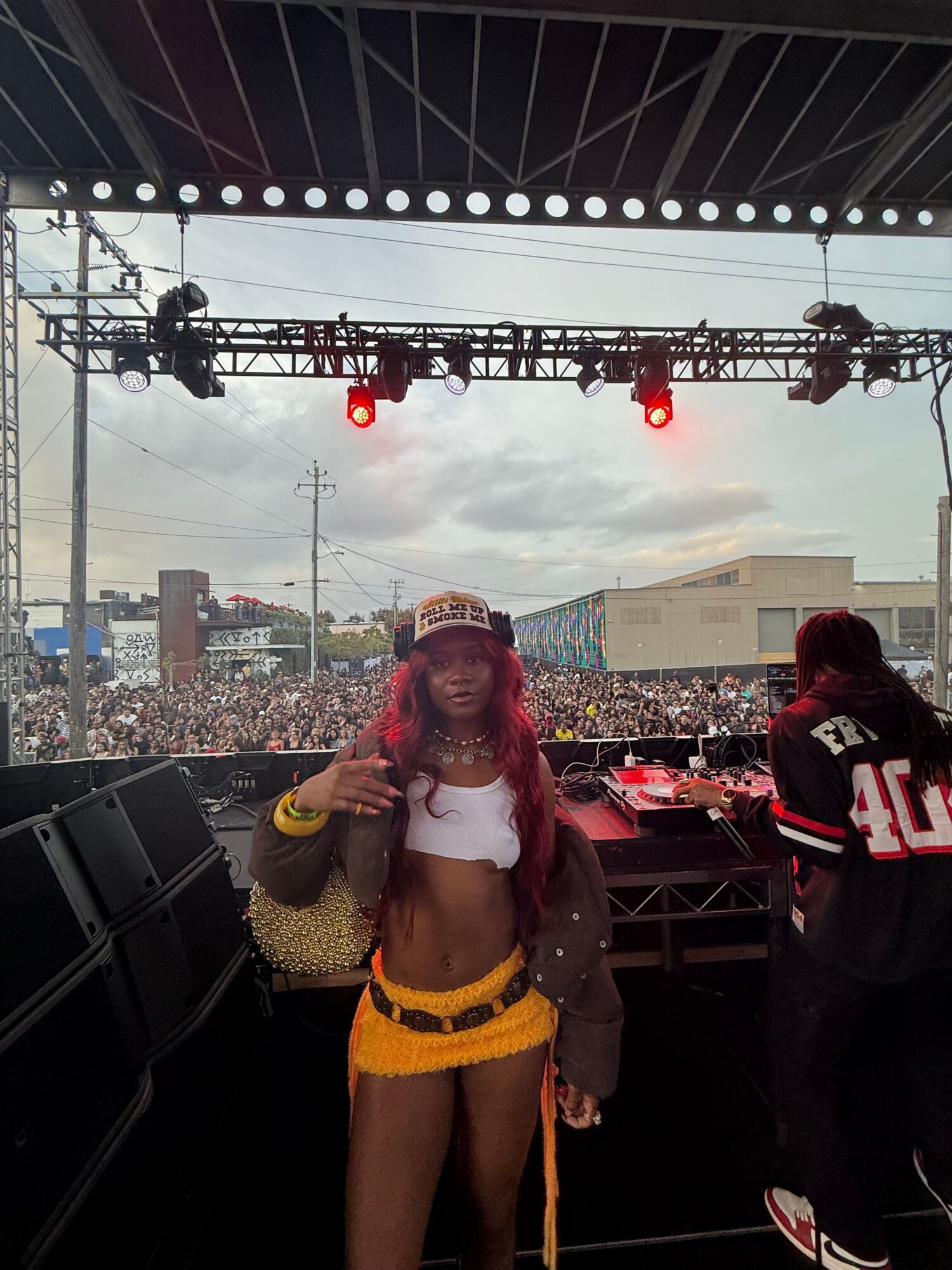 A person in a white top and yellow skirt stands onstage near a microphone, with a crowd and stage equipment behind them.